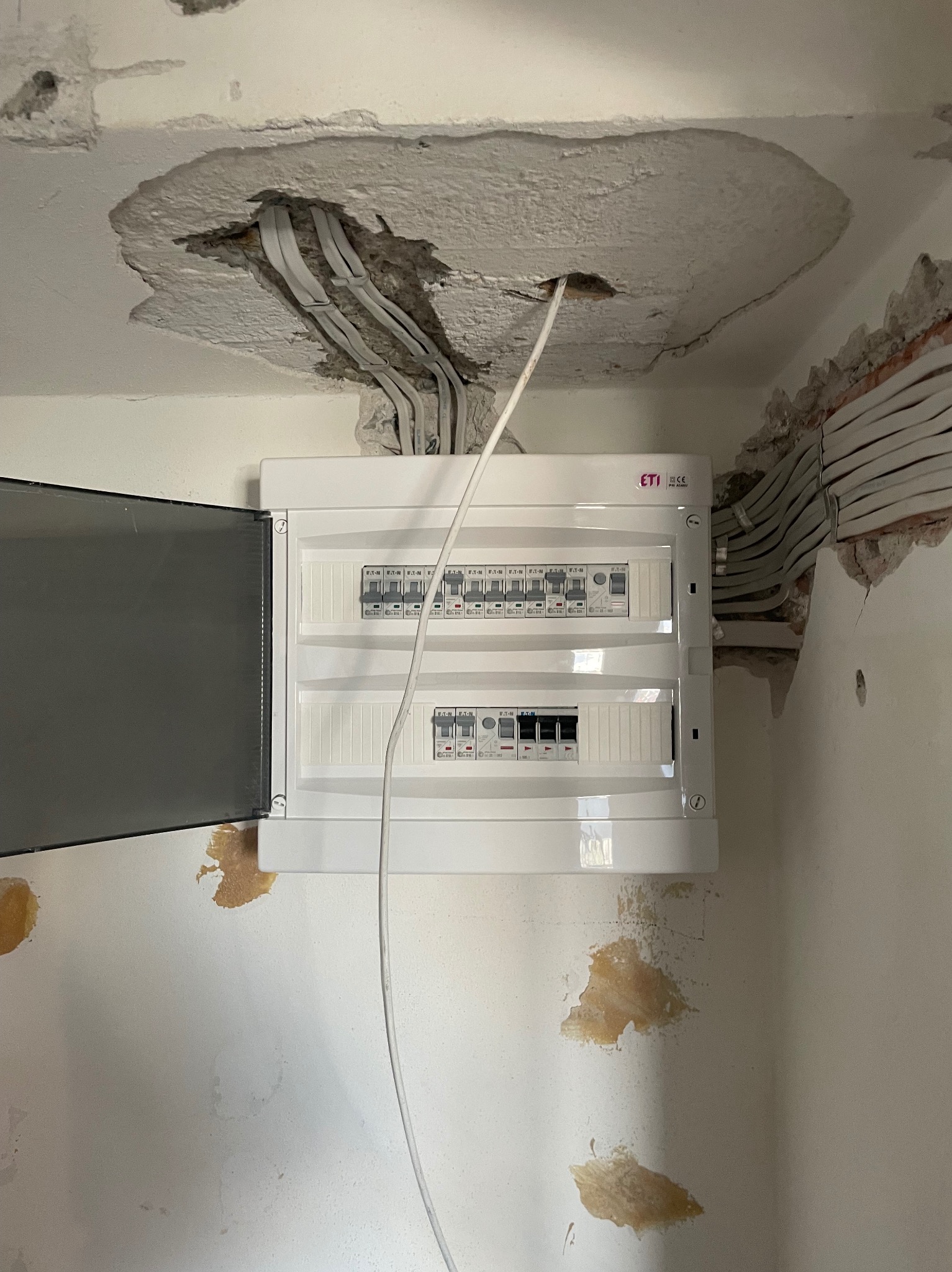 Nowa, biała skrzynka rozdzielcza ETI z otwartymi drzwiczkami, zamontowana na ścianie w trakcie remontu. Widoczne kable w suficie i ścianie.