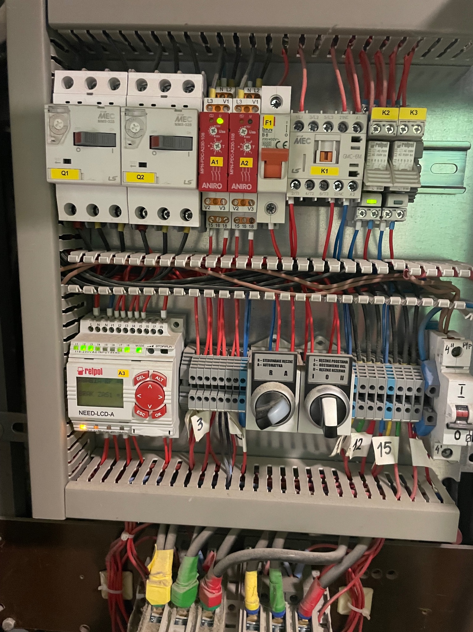 Rozdzielnica elektryczna z widocznymi elementami sterującymi, przewodami i oznaczeniami. Widoczne bezpieczniki, przekaźniki i sterownik PLC Relpol.