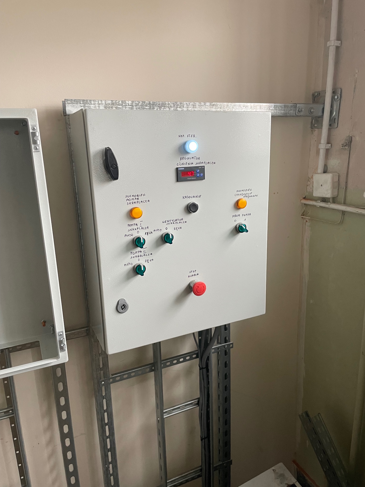 Szara szafa sterownicza z przełącznikami i wyświetlaczem, zamontowana na ścianie. Widoczne przewody i elementy instalacji. Napisy na obudowie w języku polskim.