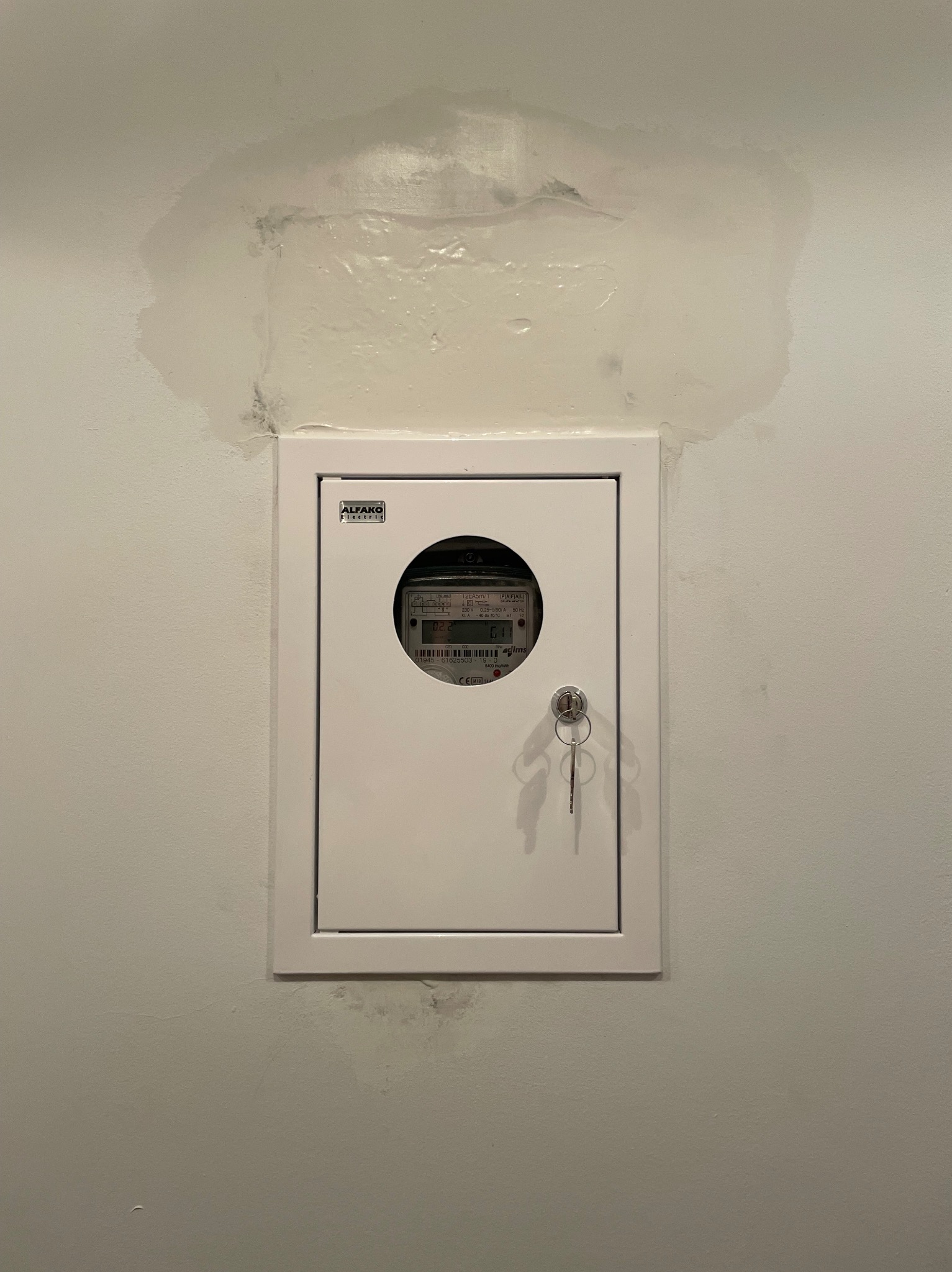 Biała skrzynka z licznikiem elektrycznym na ścianie z widocznymi śladami wilgoci. Kluczyk w zamku. Widoczna marka 'ALFAKO Electric'.
