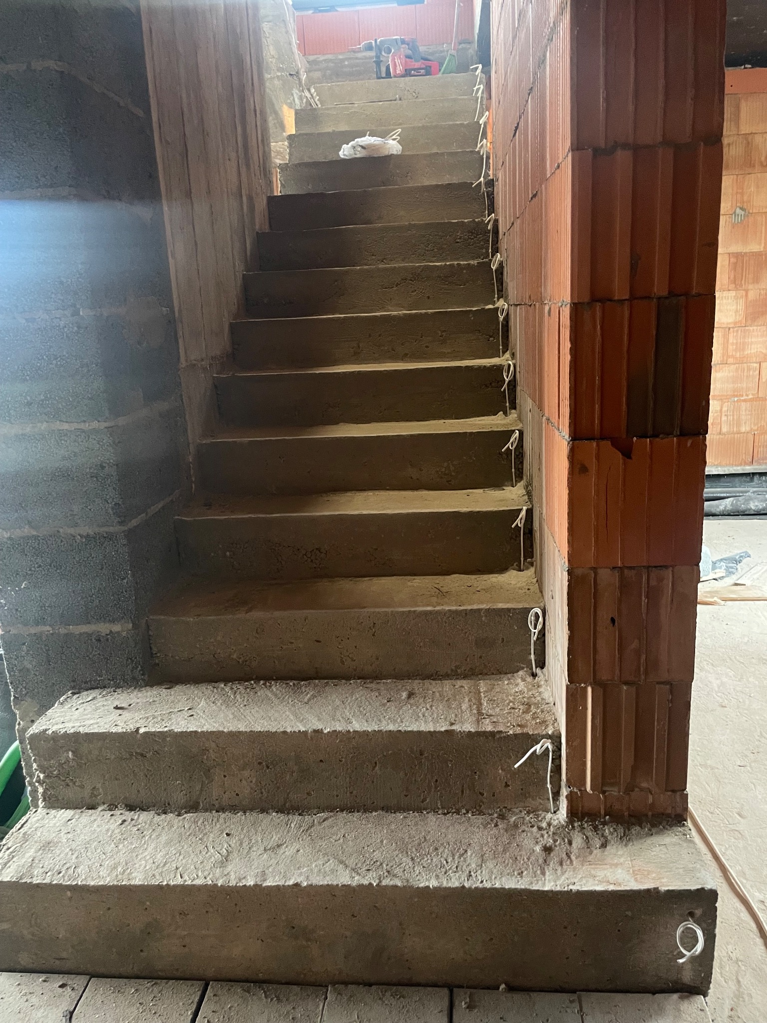 Surowe betonowe schody w trakcie budowy, z widocznymi ceglanymi filarami i wystającymi przewodami. W tle narzędzia budowlane i materiały.