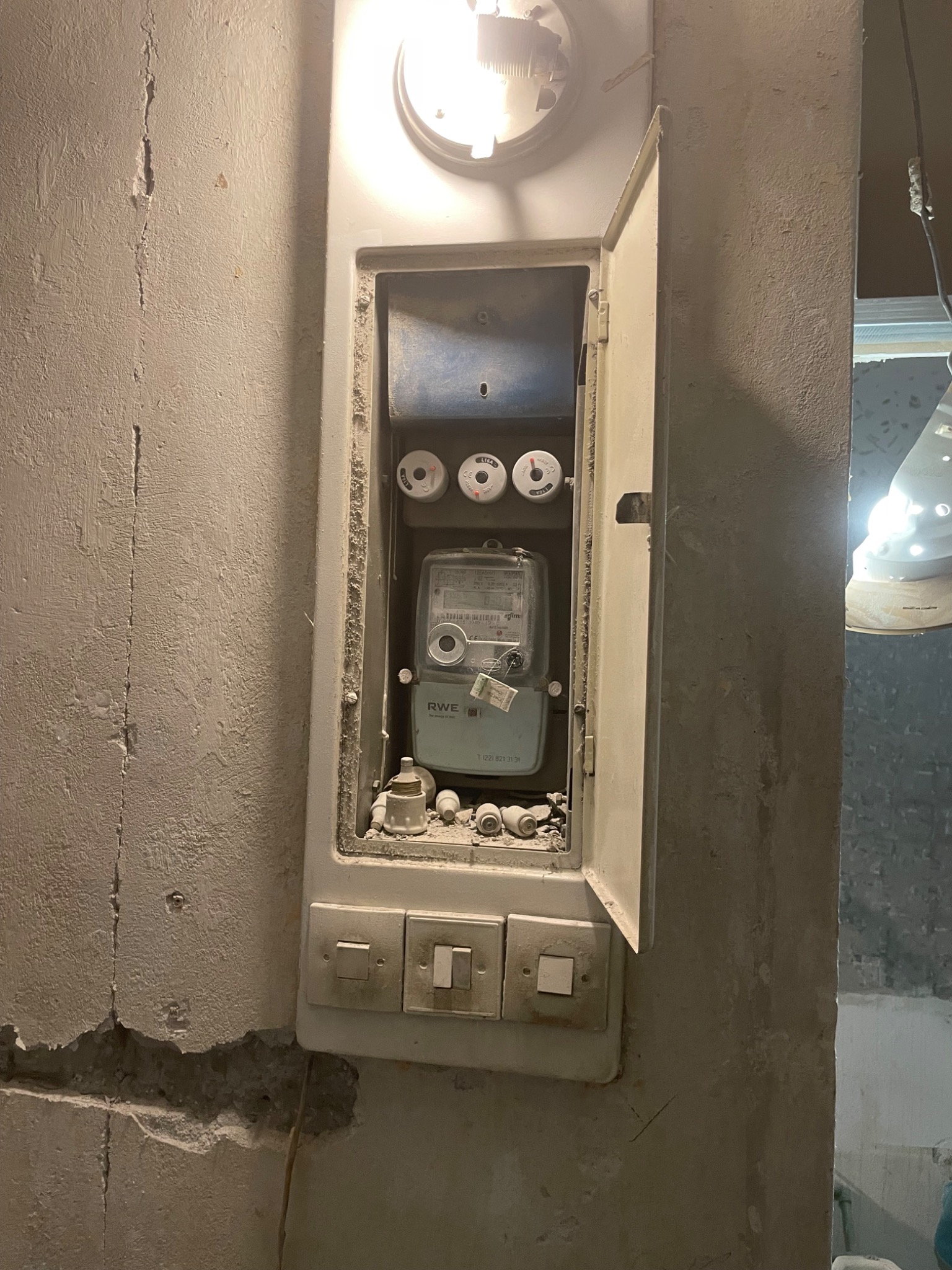 Otwarta skrzynka z licznikiem RWE i bezpiecznikami na brudnej ścianie z włącznikami. Widoczne ślady po kuciu i pęknięcia na ścianie.