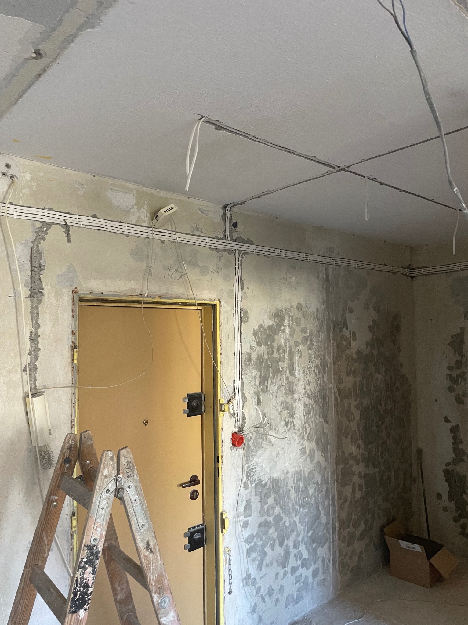 Wnętrze w trakcie remontu z widocznymi przewodami elektrycznymi ułożonymi wzdłuż ścian i sufitu. Drabina oparta o futrynę drzwi. Surowe ściany przygotowane do wykończenia.