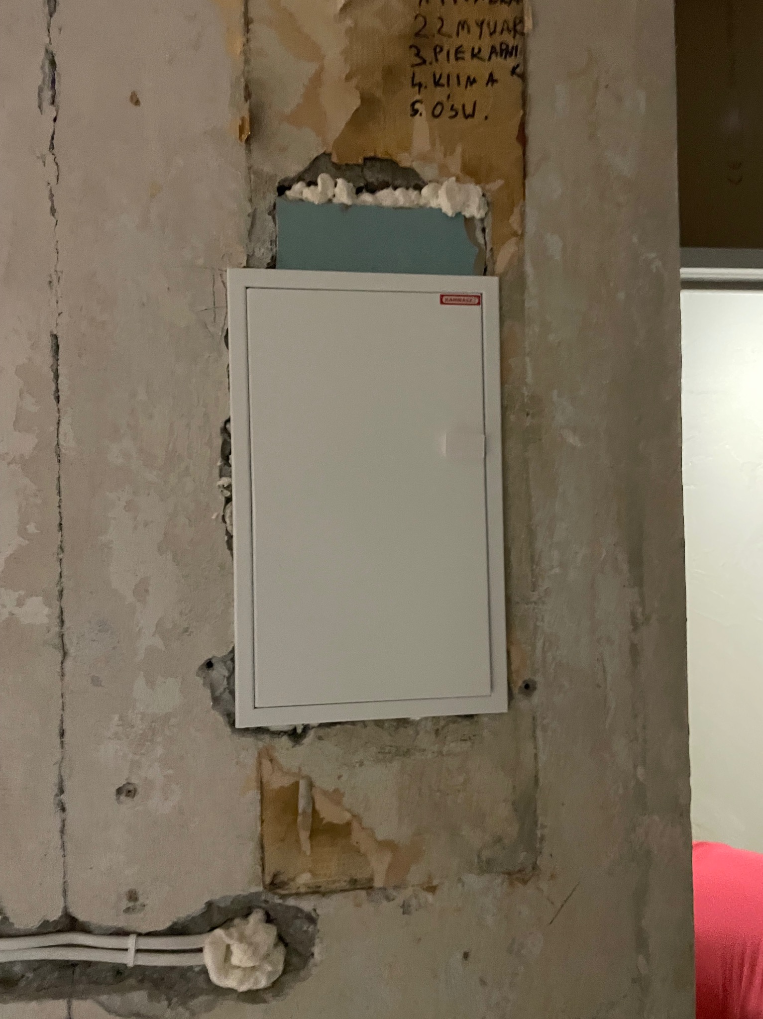 Biała skrzynka elektryczna w surowej ścianie z widocznymi przewodami i notatkami. Etap prac instalacyjnych przed wykończeniem.