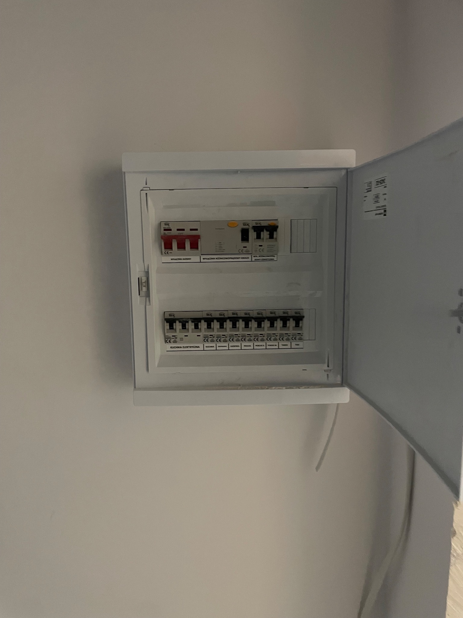 Biała skrzynka rozdzielcza z bezpiecznikami na ścianie, otwarte drzwiczki ukazują wnętrze z wyłącznikami. Widoczne oznaczenia i certyfikaty na urządzeniach.