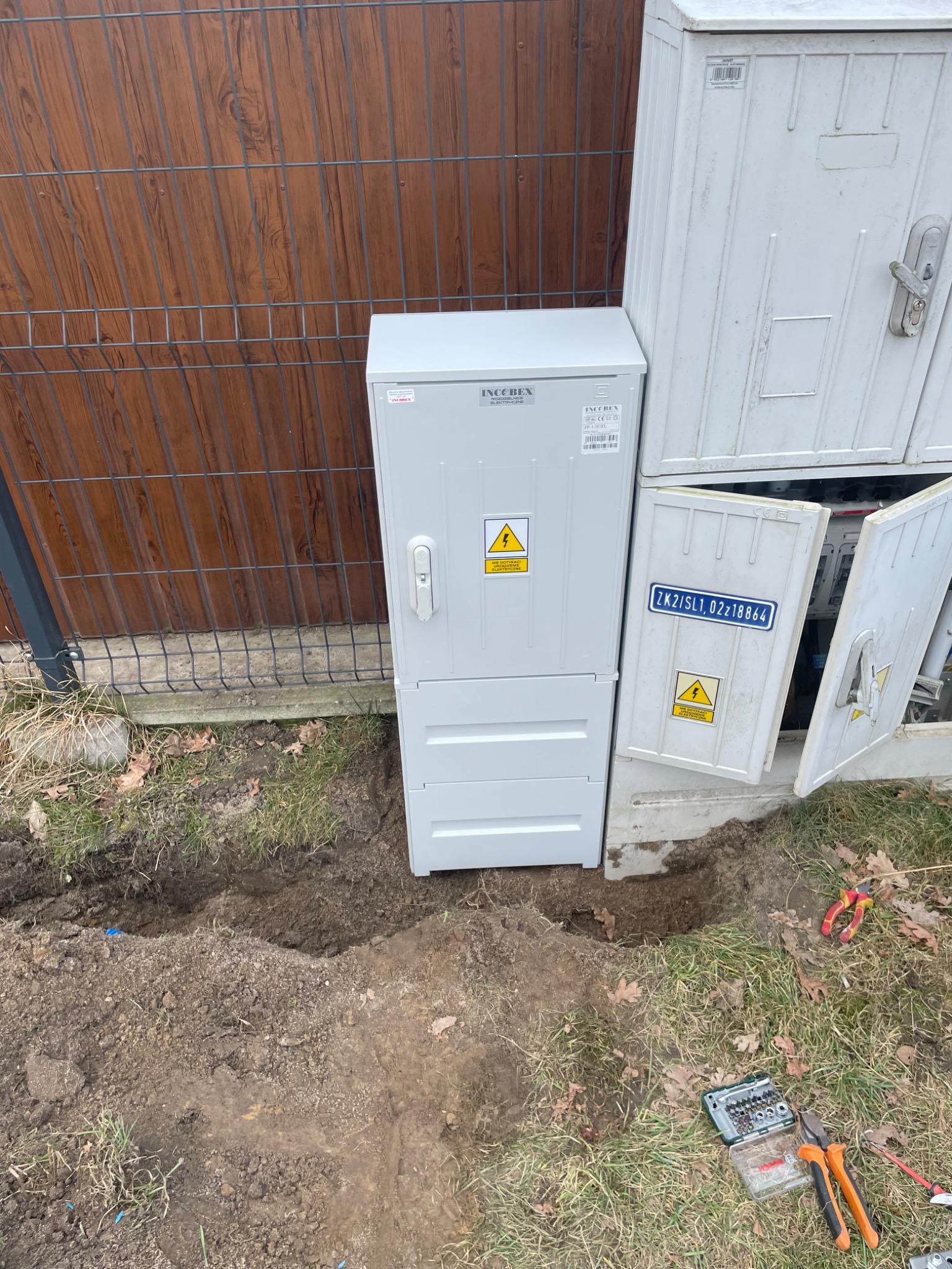 Nowa, szara skrzynka elektryczna IncoBex obok starej, otwartej skrzynki. Wykopany rów w ziemi sugeruje prace przyłączeniowe. Widoczne narzędzia.