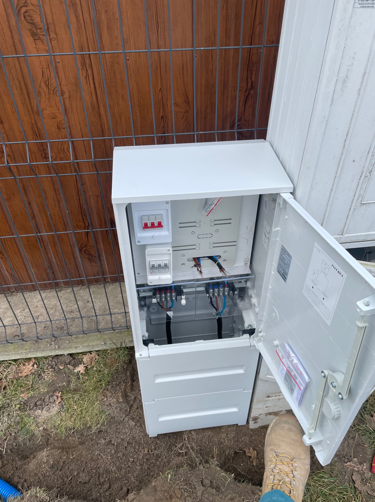 Biała skrzynka elektryczna z otwartymi drzwiczkami, widoczne bezpieczniki i przewody, umieszczona na zewnątrz, obok ogrodzenia z siatki i drewnianego płotu.