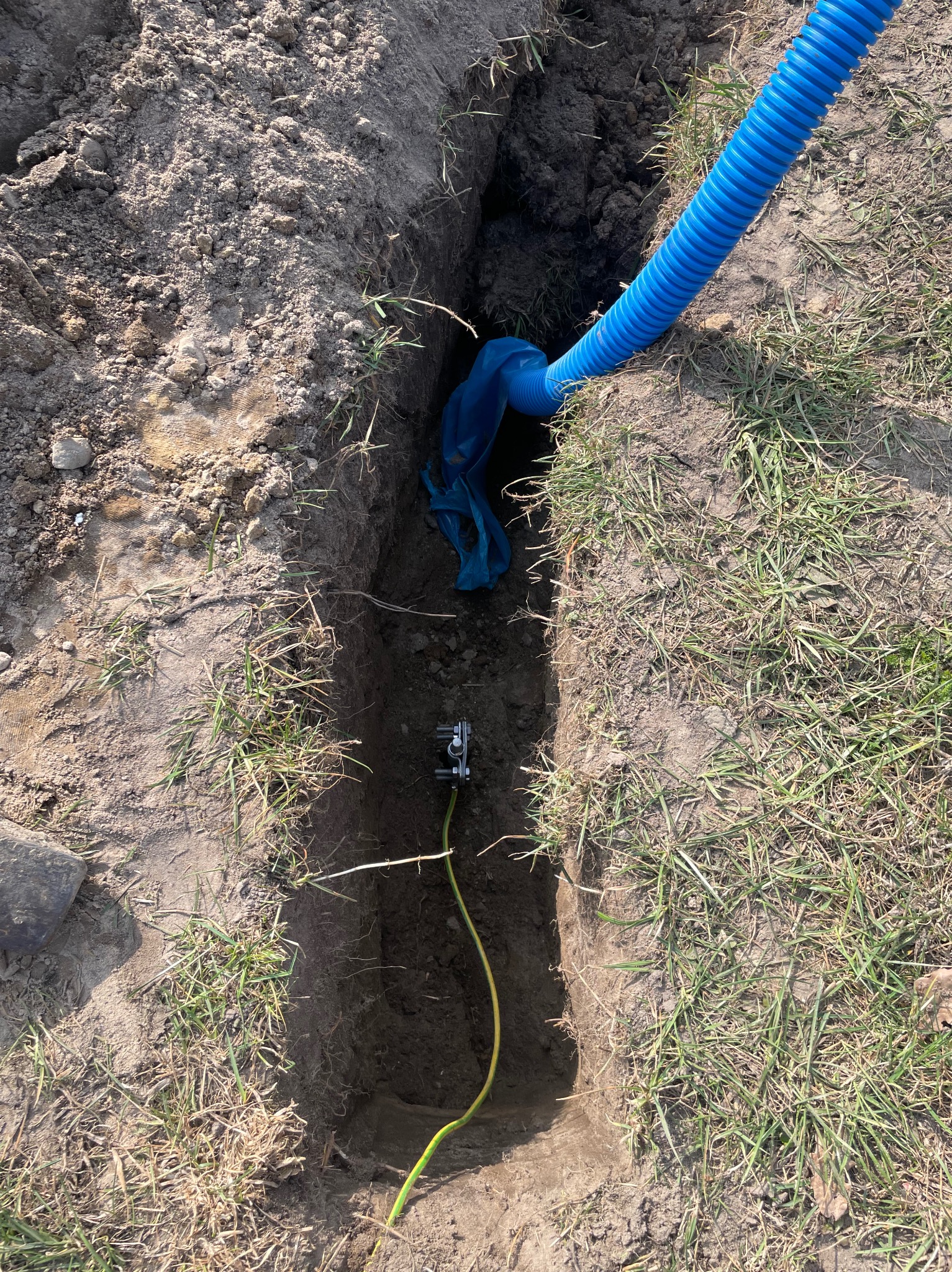 Wkopana w ziemię niebieska rura karbowana z kablem uziemiającym, widoczne złącze. Grunt piaszczysty z trawą. Ujęcie z góry, w dziennym świetle.