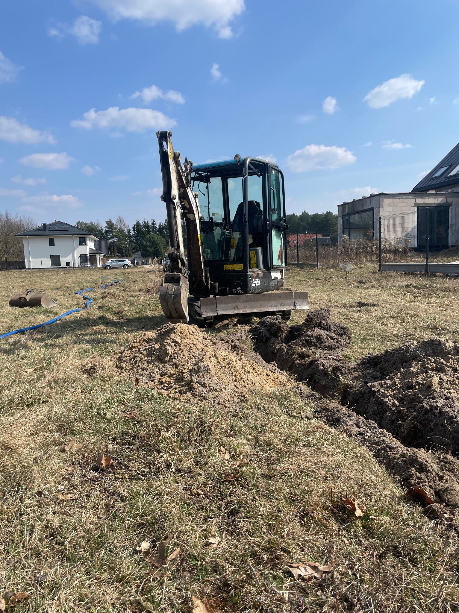 Mini koparka wykopuje rów pod instalację elektryczną w Warszawie. W tle domy jednorodzinne i błękitne niebo. Widoczne niebieskie rury.