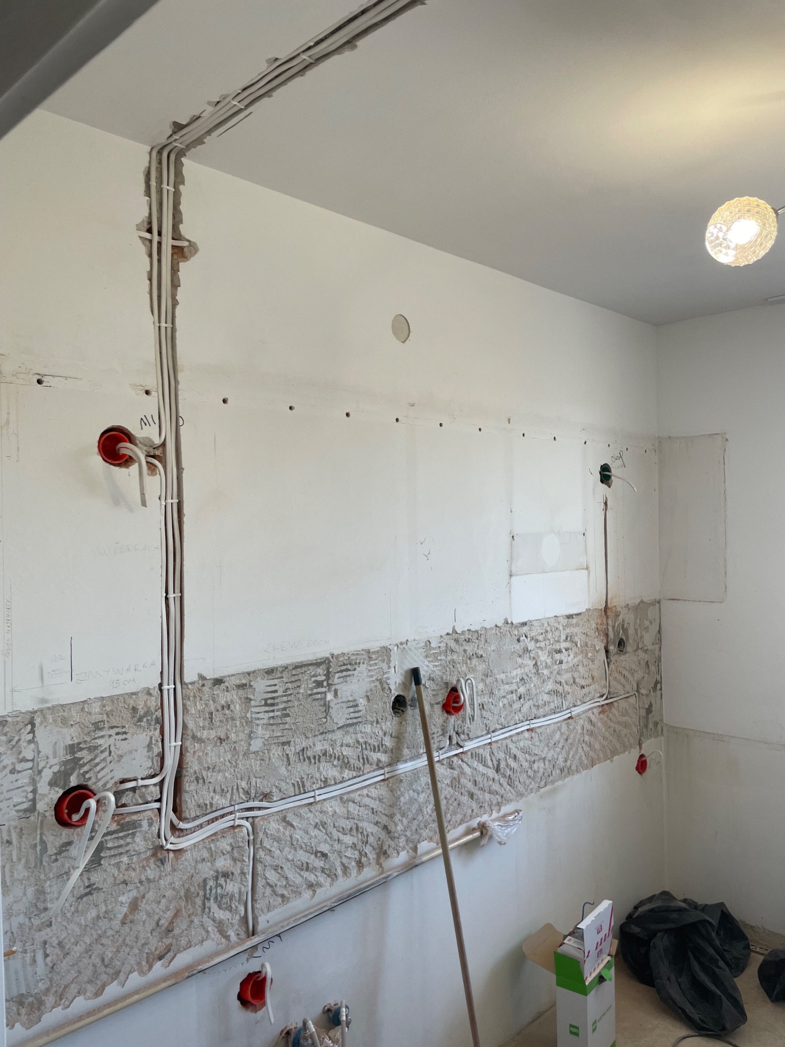 Białe przewody elektryczne w bruzdach na ścianie w trakcie montażu, widoczne puszki instalacyjne i fragment instalacji oświetleniowej pod sufitem.