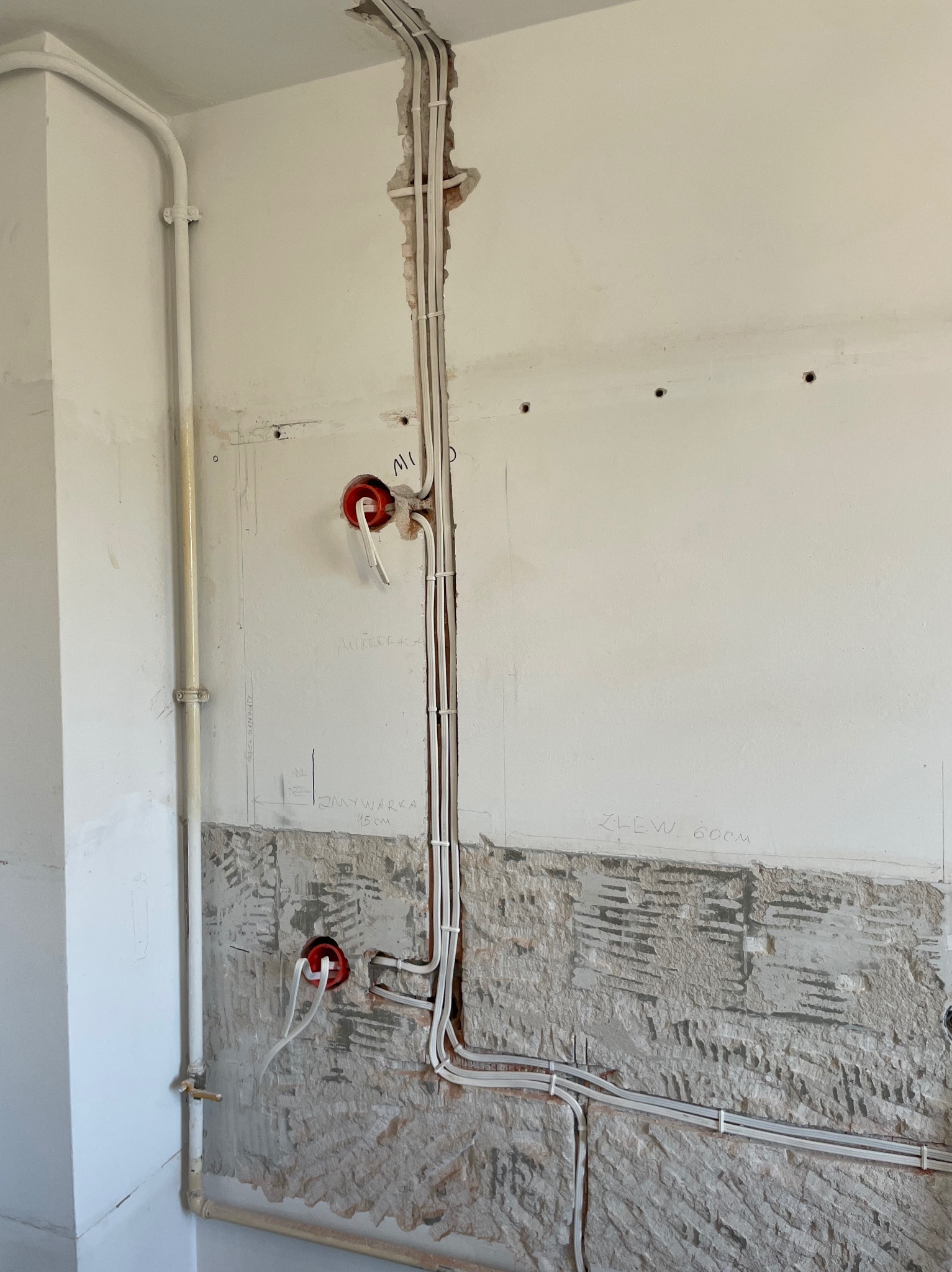 Białe kable elektryczne prowadzone wzdłuż ściany z widocznymi puszkami instalacyjnymi i fragmentami odsłoniętej cegły, zarysowane plany instalacji na ścianie.