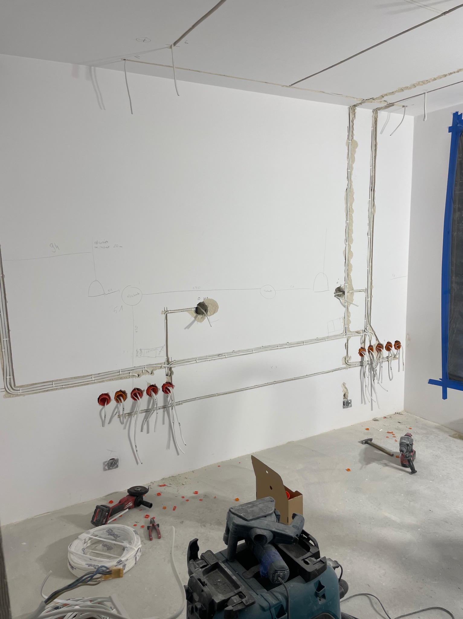 Rozprowadzone przewody elektryczne w bruzdach na ścianie, przygotowane pod gniazdka i oświetlenie. Widoczne narzędzia i materiały budowlane na podłodze w surowym wnętrzu.