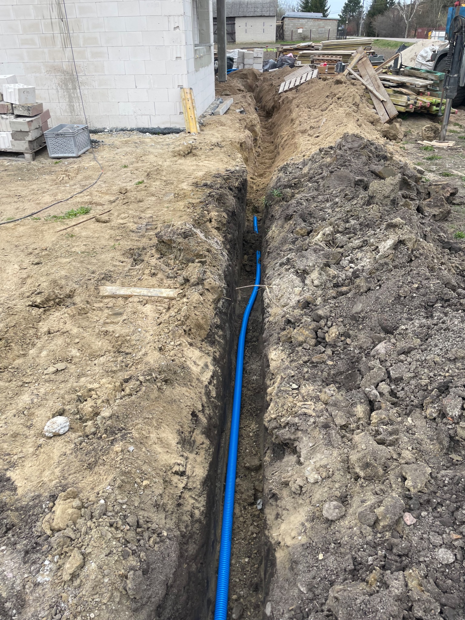 Niebieski karbowany przewód w wykopie ziemnym, prawdopodobnie do instalacji elektrycznej, na tle budowy domu z białego bloczka.