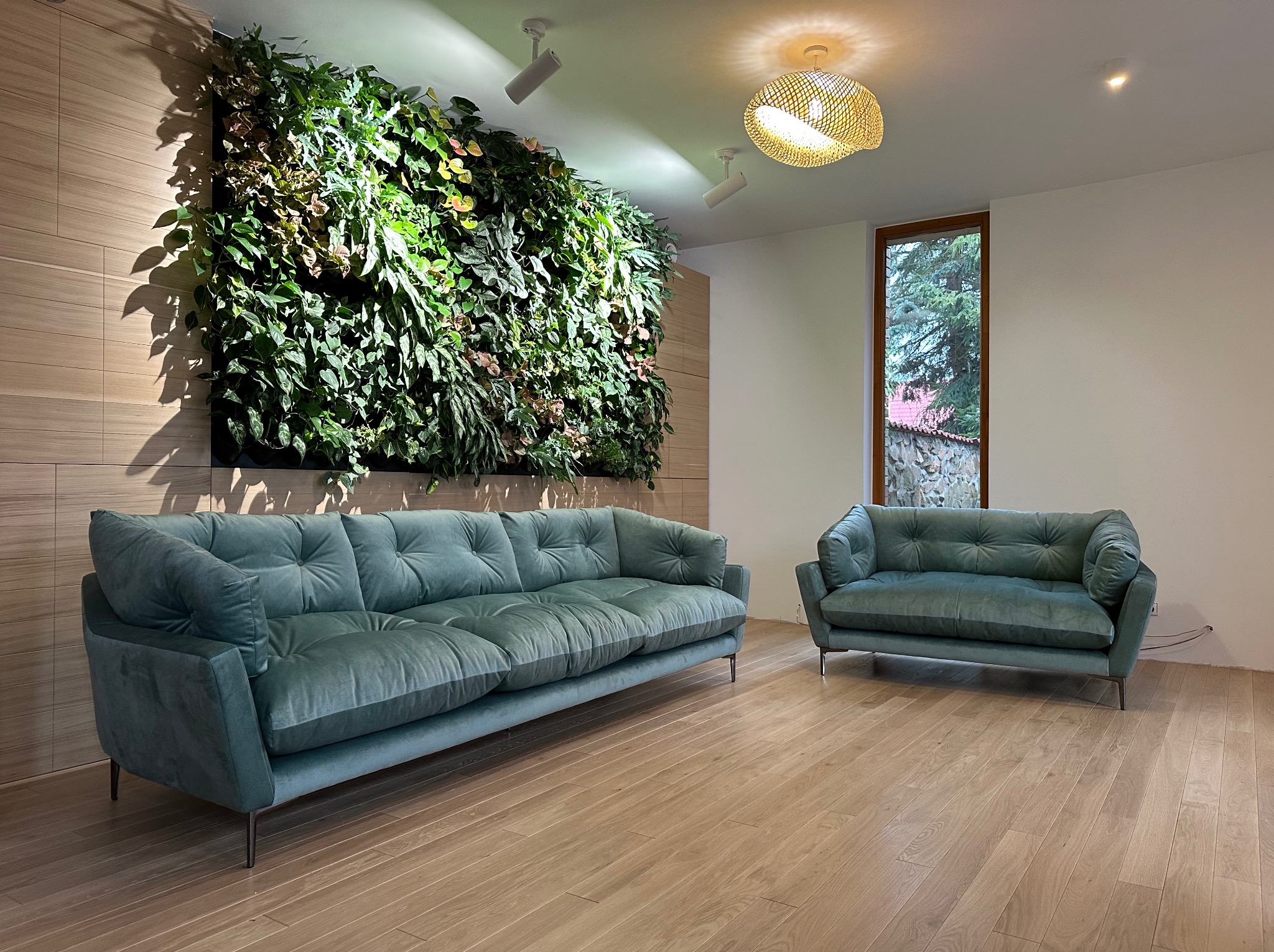 Turkusowy, pikowany zestaw wypoczynkowy: sofa trzyosobowa i fotel, na metalowych nóżkach, na tle ściany z zielenią w nowoczesnym wnętrzu.