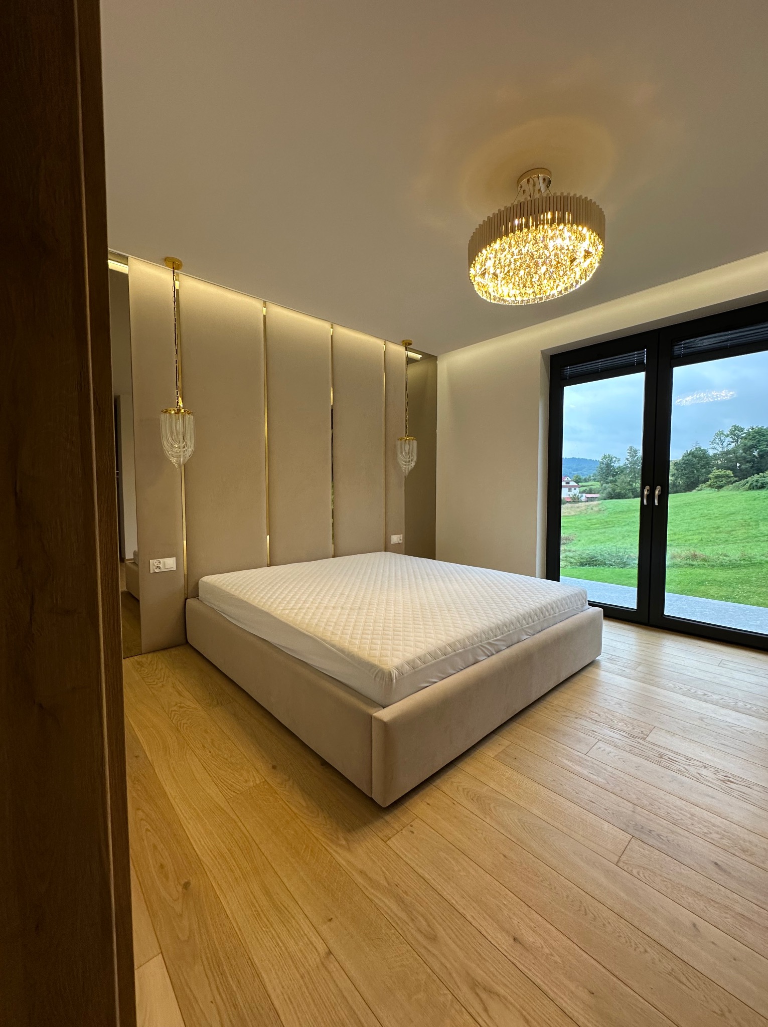 Nowoczesna sypialnia z beżowym łóżkiem kontynentalnym, tapicerowanym wezgłowiem z pionowymi, złotymi akcentami i widokiem na zielony krajobraz przez duże okno.