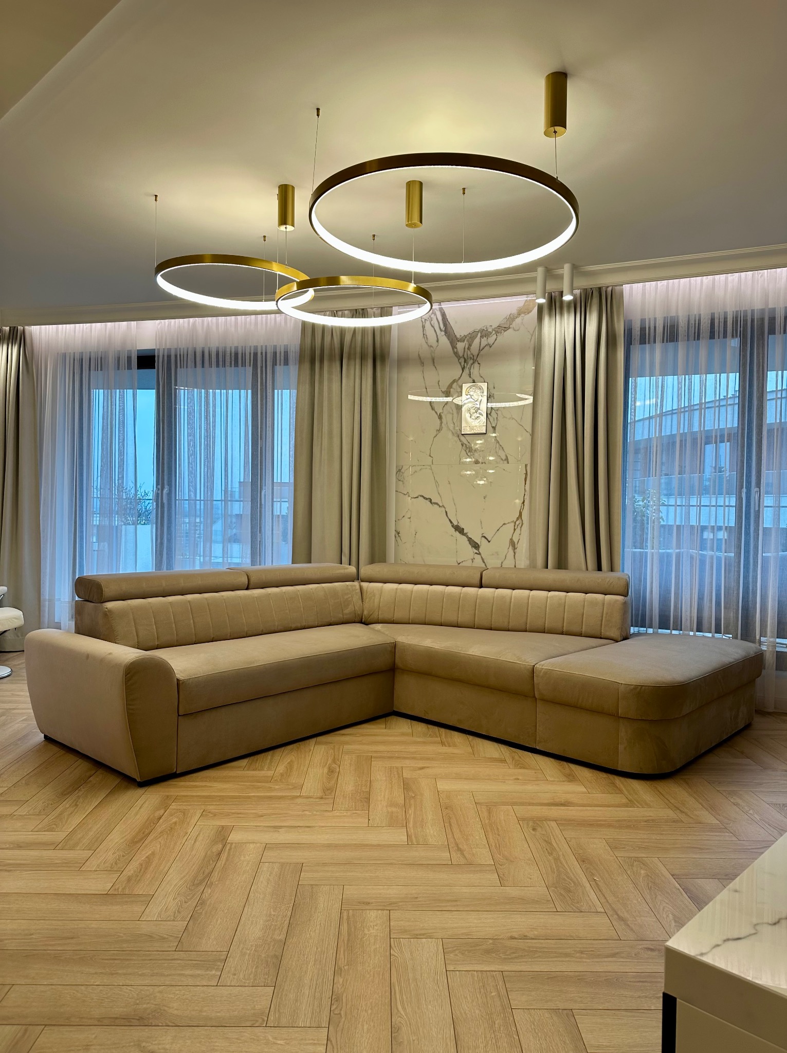 Beżowa narożna sofa tapicerowana w nowoczesnym salonie z parkietem w jodełkę, oświetlona trzema okrągłymi lampami wiszącymi z elementami w kolorze złota.