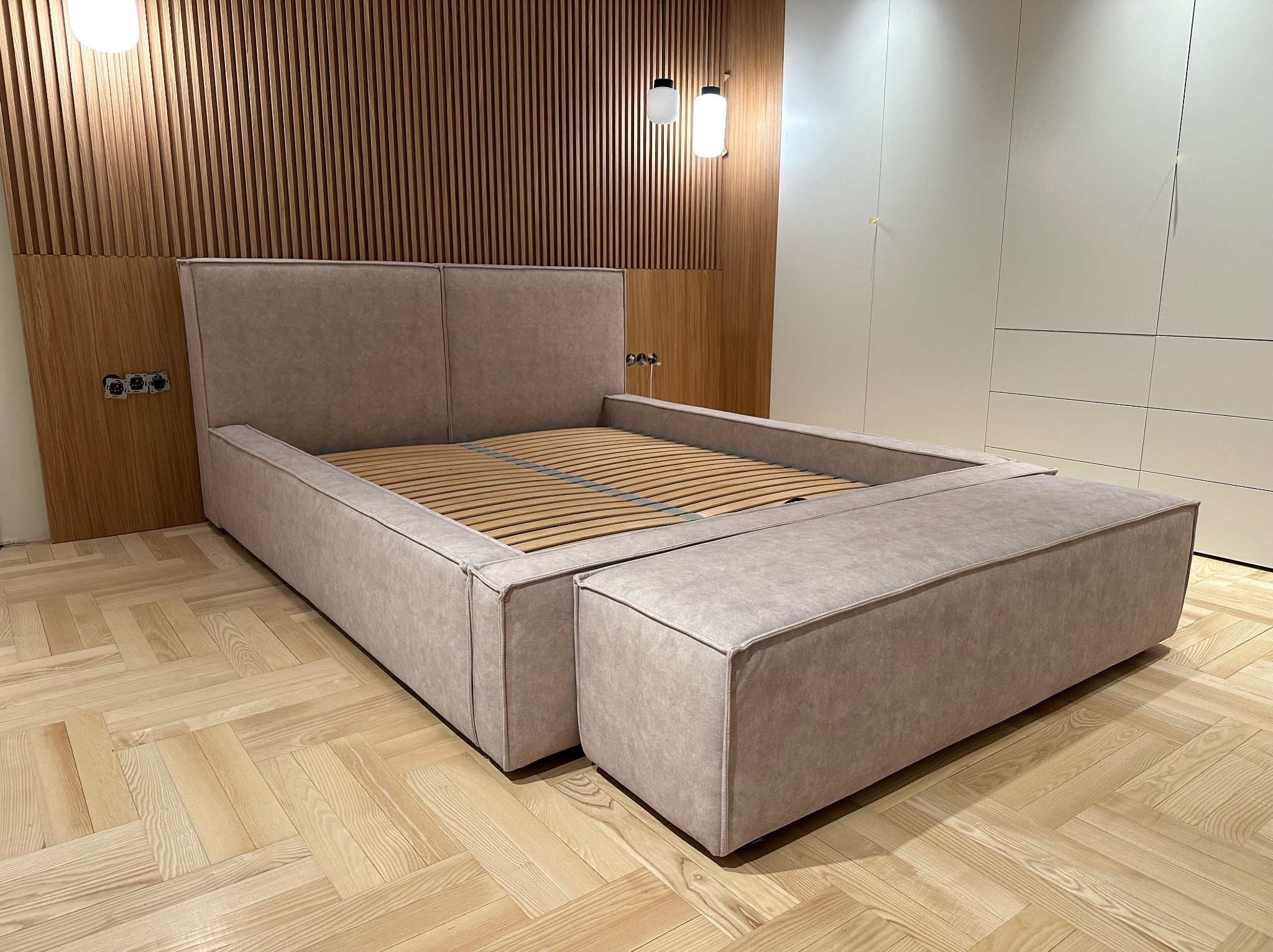 Beżowe łóżko tapicerowane z drewnianym stelażem i dopasowaną pufą, na parkiecie w jodełkę, z drewnianą ścianą z listew i białymi szafami w tle.