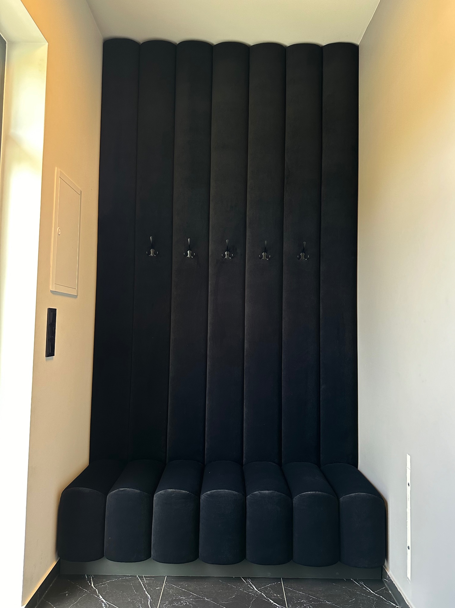 Czarne, welurowe panele ścienne z wieszakami i siedziskiem w przedpokoju, marmurowa podłoga.