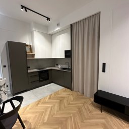 SOK&MAN - Nowoczesna kuchnia z szarymi szafkami, białymi górnymi szafkami i parkietem ułożonym w jodełkę; widoczny fragment salonu z czarną szafką RTV.