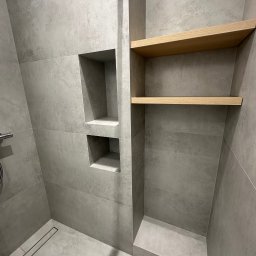 SOK&MAN - Wykończona łazienka z szarymi płytkami imitującymi beton, drewnianymi półkami w zabudowie i dwoma niszami w ścianie obok kabiny prysznicowej z odpływem liniowym.