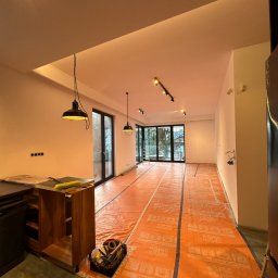 SOK&MAN - Przygotowanie do układania paneli w przestronnym salonie z dużymi oknami, podłoga zabezpieczona pomarańczową folią ochronną, widoczny zestaw narzędzi i częściowo zmontowana szafka kuchenna.