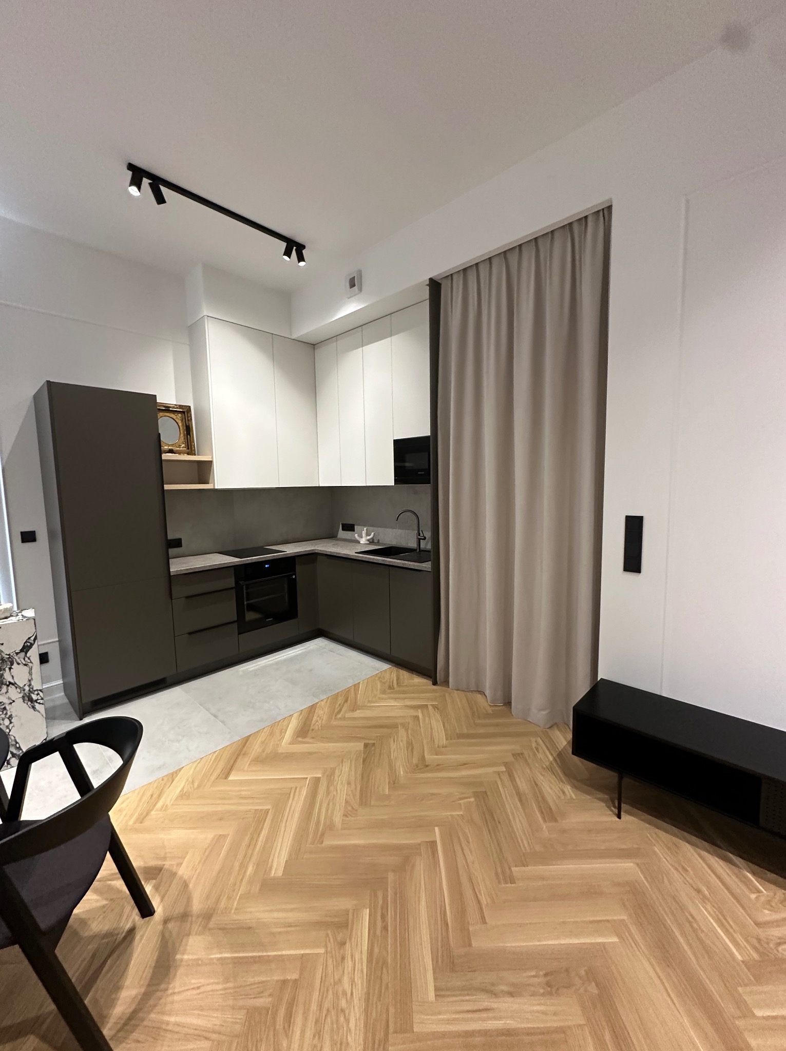 Nowoczesna kuchnia z szarymi szafkami, białymi górnymi szafkami i parkietem ułożonym w jodełkę; widoczny fragment salonu z czarną szafką RTV.