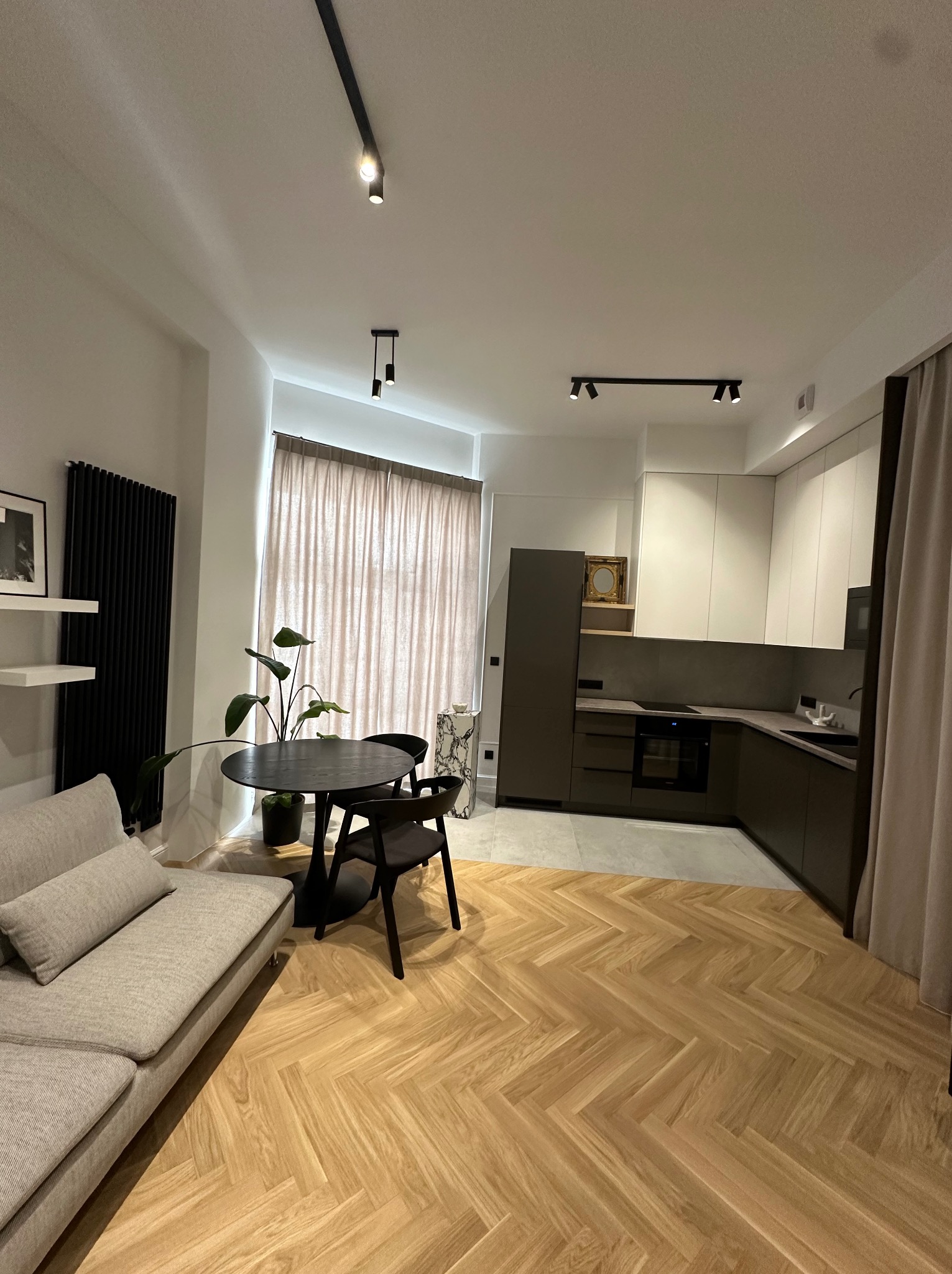 Nowoczesne wnętrze mieszkania z parkietem w jodełkę, szarą sofą, okrągłym stołem z rośliną, czarnym grzejnikiem i zabudową kuchenną w kolorach bieli i grafitu.