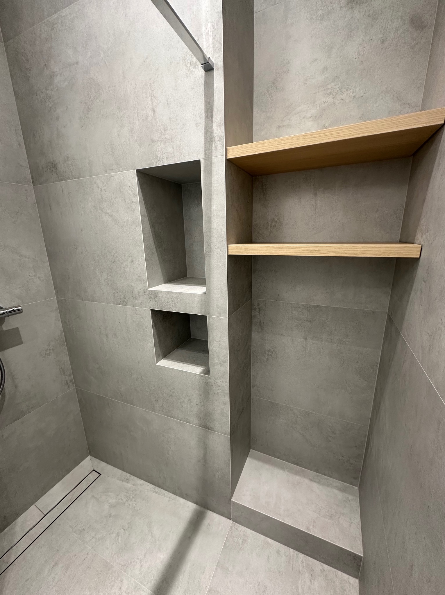 Wykończona łazienka z szarymi płytkami imitującymi beton, drewnianymi półkami w zabudowie i dwoma niszami w ścianie obok kabiny prysznicowej z odpływem liniowym.