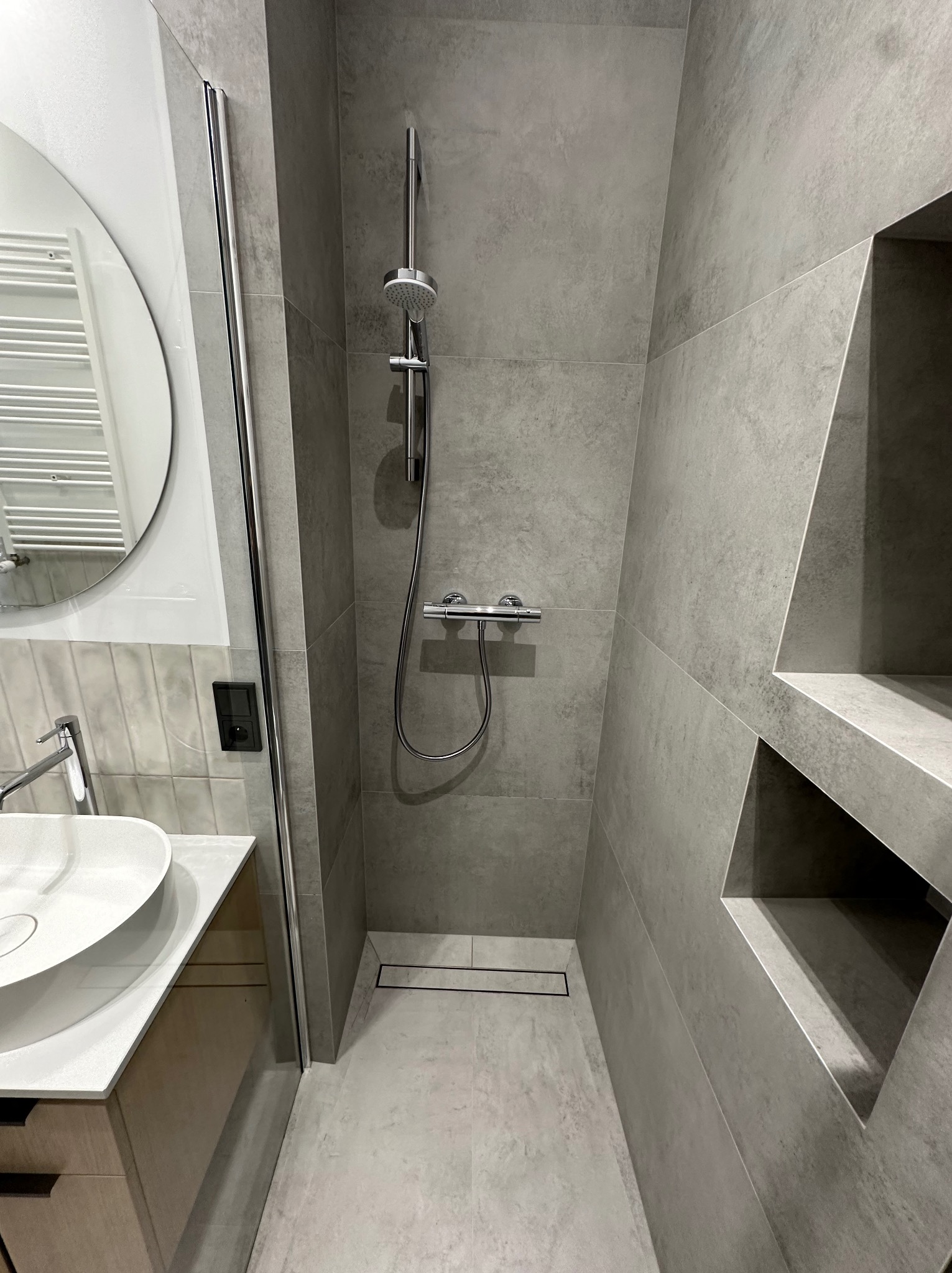 Nowoczesna łazienka w odcieniach szarości z kabiną prysznicową typu walk-in, odpływem liniowym i wnękami w ścianie.