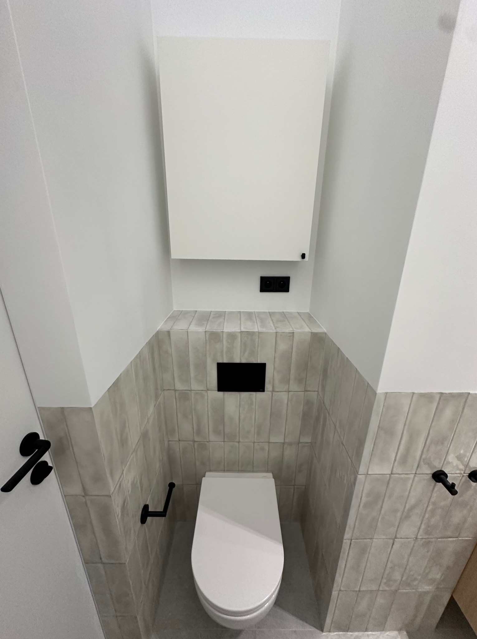 Nowoczesna toaleta w łazience z szarymi płytkami ułożonymi pionowo, czarnymi akcentami armatury i ukrytą spłuczką.