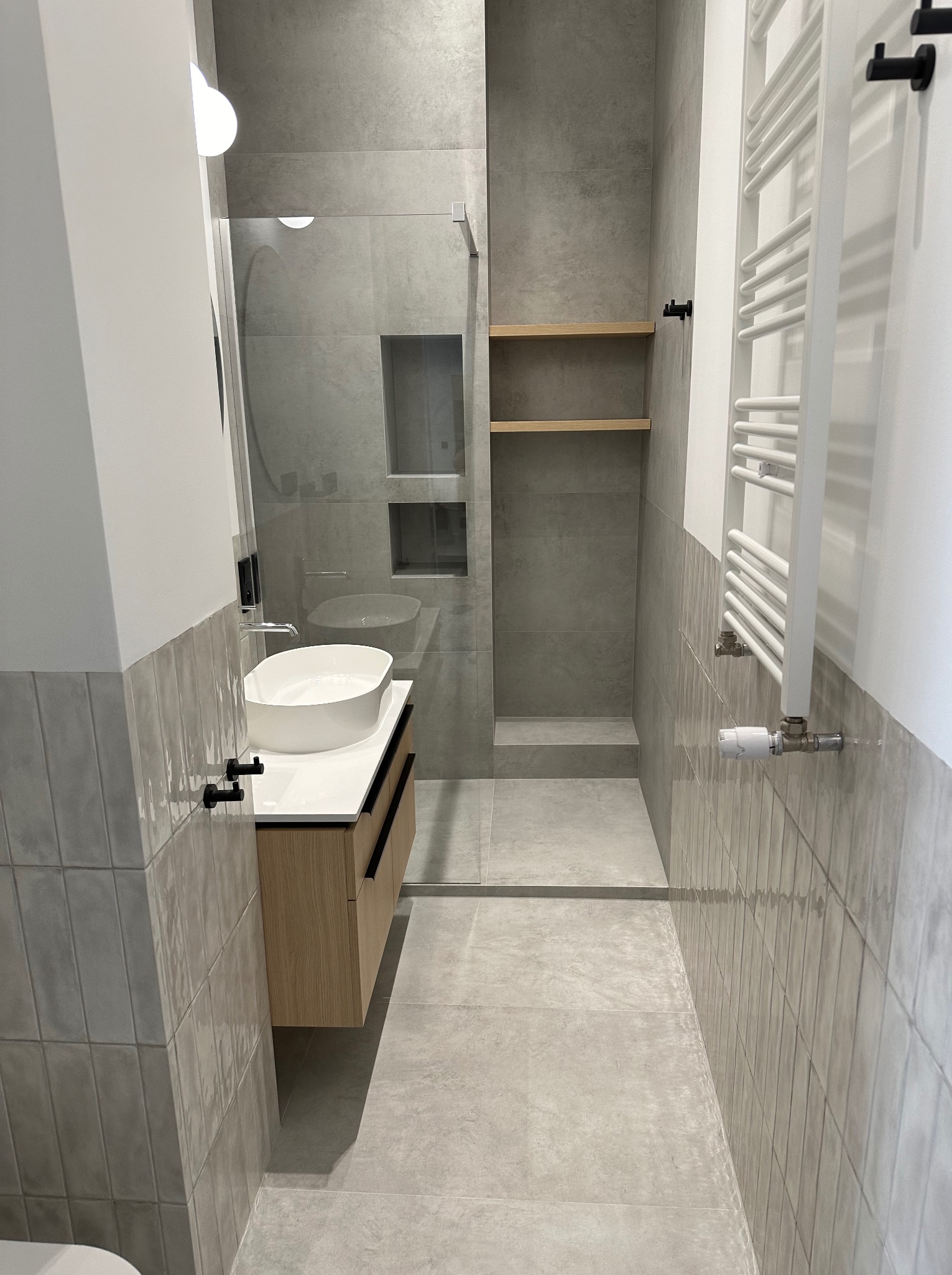 Nowoczesna łazienka w odcieniach szarości z kabiną prysznicową typu walk-in, umywalką nablatową na drewnianej szafce i grzejnikiem drabinkowym.