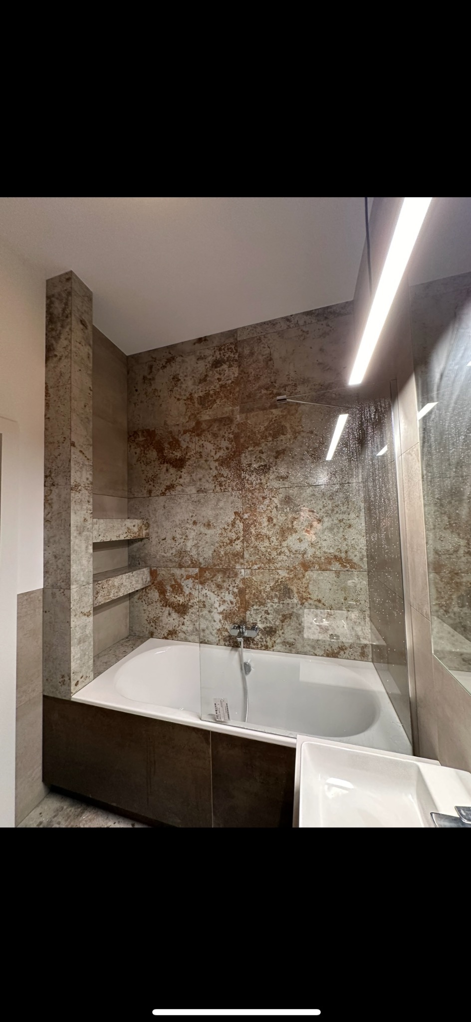 Nowoczesna łazienka z wanną, szklanym panelem prysznicowym i płytkami imitującymi rdzę, oświetlenie LED w górnej części.