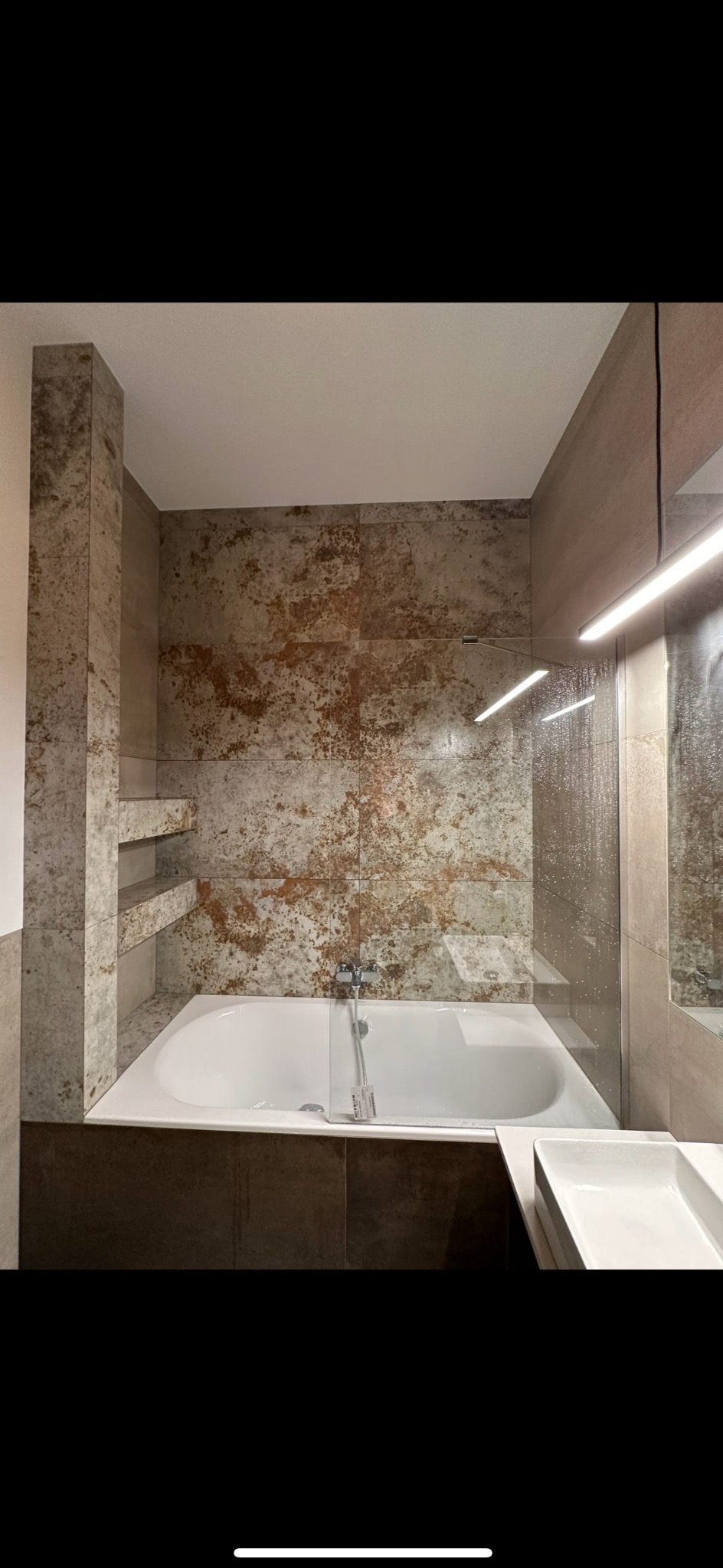 Wanna z parawanem, obłożona płytkami imitującymi rdzawy kamień, z wnękami na kosmetyki i szklanym parawanem nawannowym.