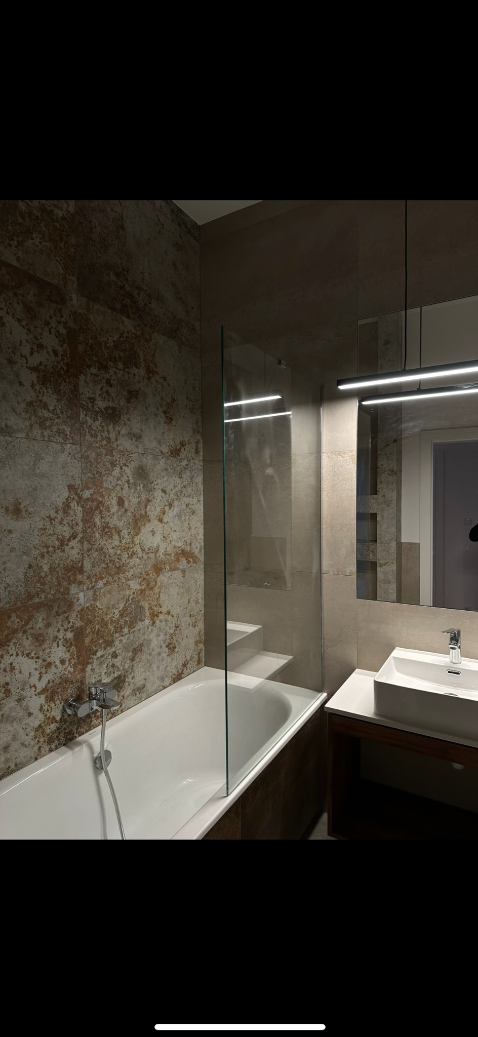 Nowoczesna łazienka z wanną obudowaną płytkami imitującymi rdzę, szklanym parawanem nawannowym i minimalistyczną umywalką nablatową z szafką z ciemnego drewna.