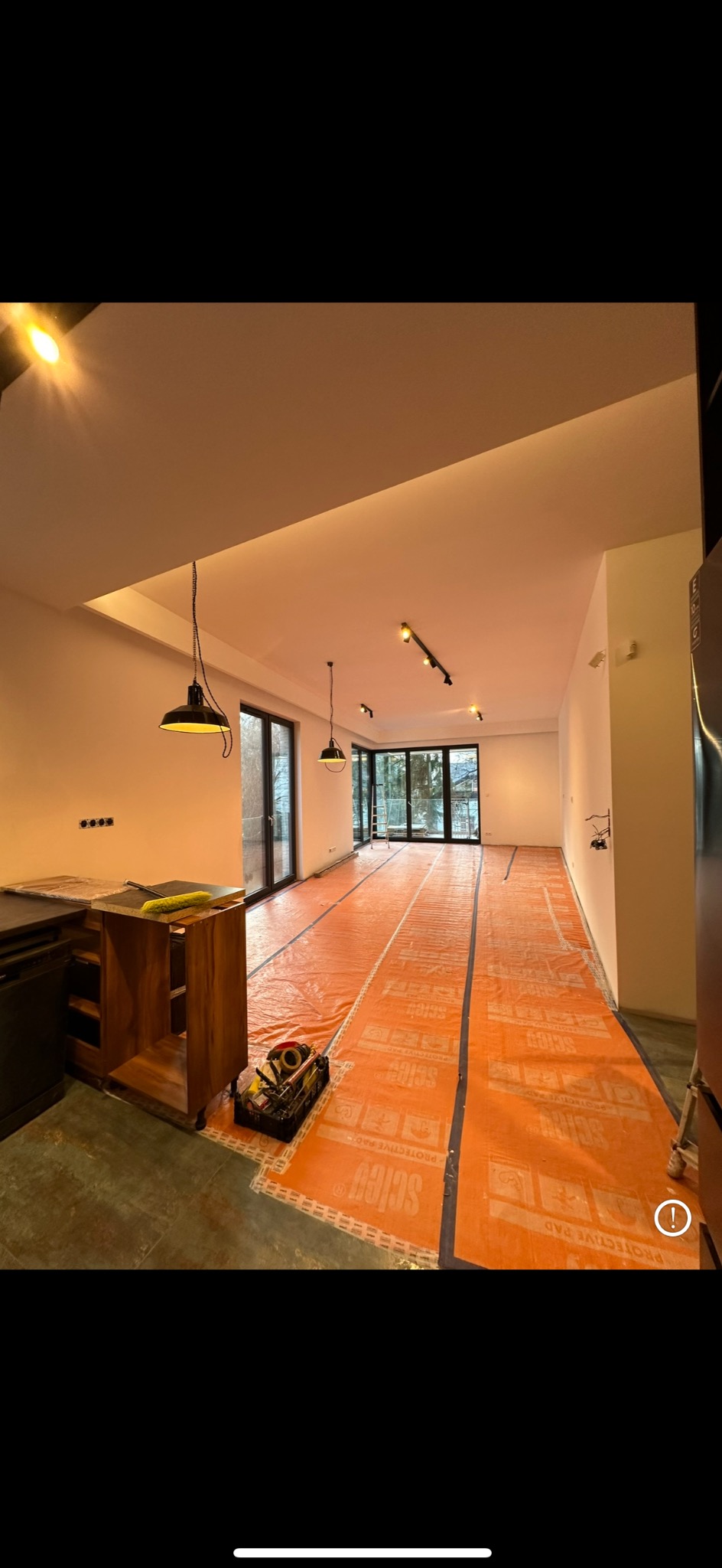 Przygotowanie do układania paneli w przestronnym salonie z dużymi oknami, podłoga zabezpieczona pomarańczową folią ochronną, widoczny zestaw narzędzi i częściowo zmontowana szafka kuchenna.