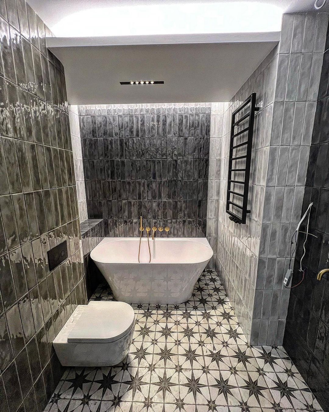 Nowoczesna łazienka z ciemnymi, błyszczącymi płytkami imitującymi cegłę, białą wolnostojącą wanną z złotą armaturą, podwieszaną toaletą i podłogą w gwiazdy.