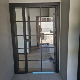 MPSMONT PIOTR KOZAKIEWICZ - Nowoczesne, przeszklone drzwi wewnętrzne z czarną ramą i podziałami, widoczne z bliska. W tle widoczne kolejne pomieszczenia i fragment ulicy.