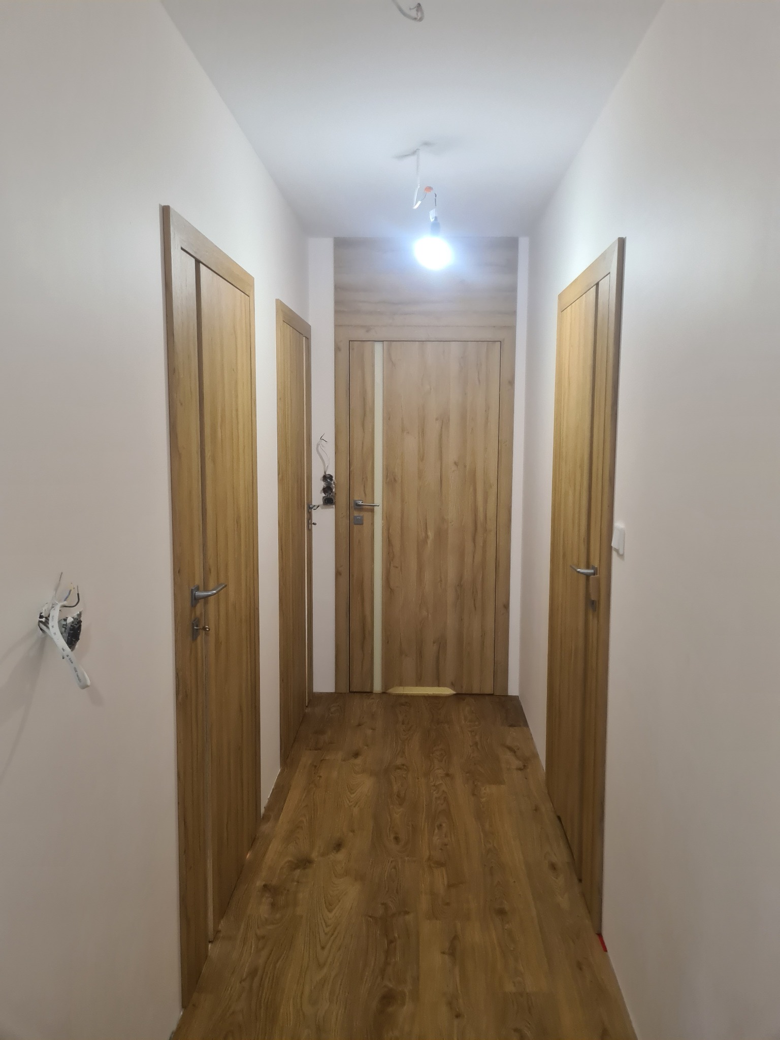Korytarz z trzema drewnianymi drzwiami o jasnej tonacji, oświetlony żarówką. Widoczne wystające kable elektryczne na ścianie, drewniana podłoga w odcieniach brązu.