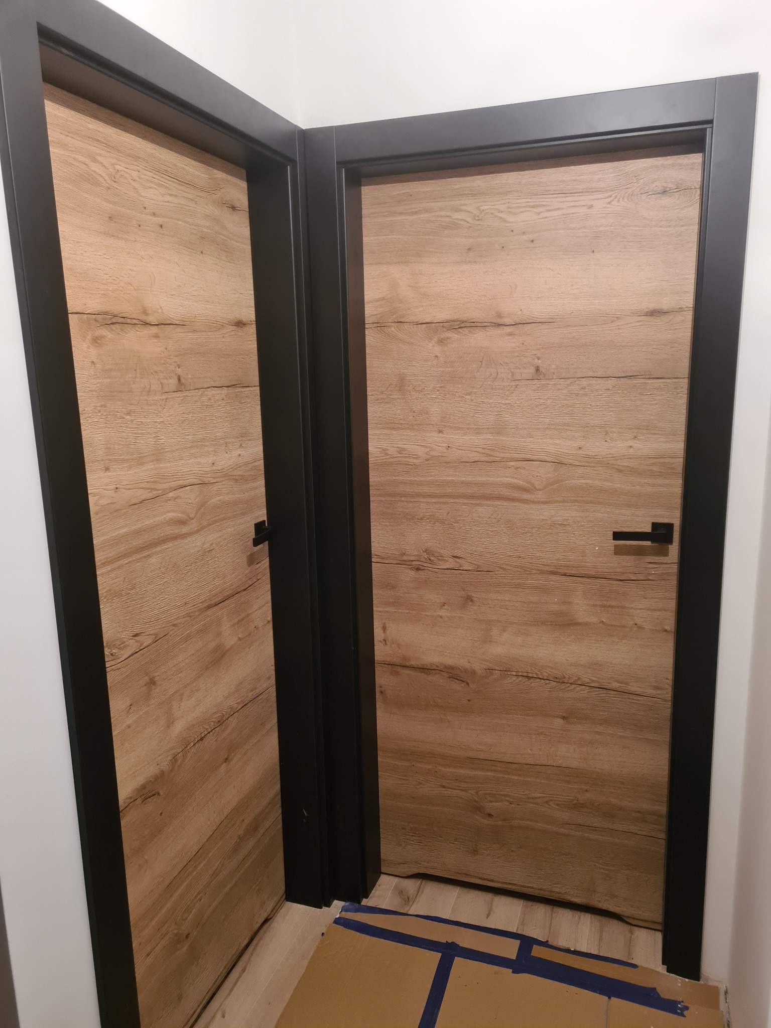 Dwa nowoczesne, drewnopodobne drzwi z czarnymi ościeżnicami, zamontowane w narożniku pomieszczenia. Podłoga zabezpieczona kartonem.