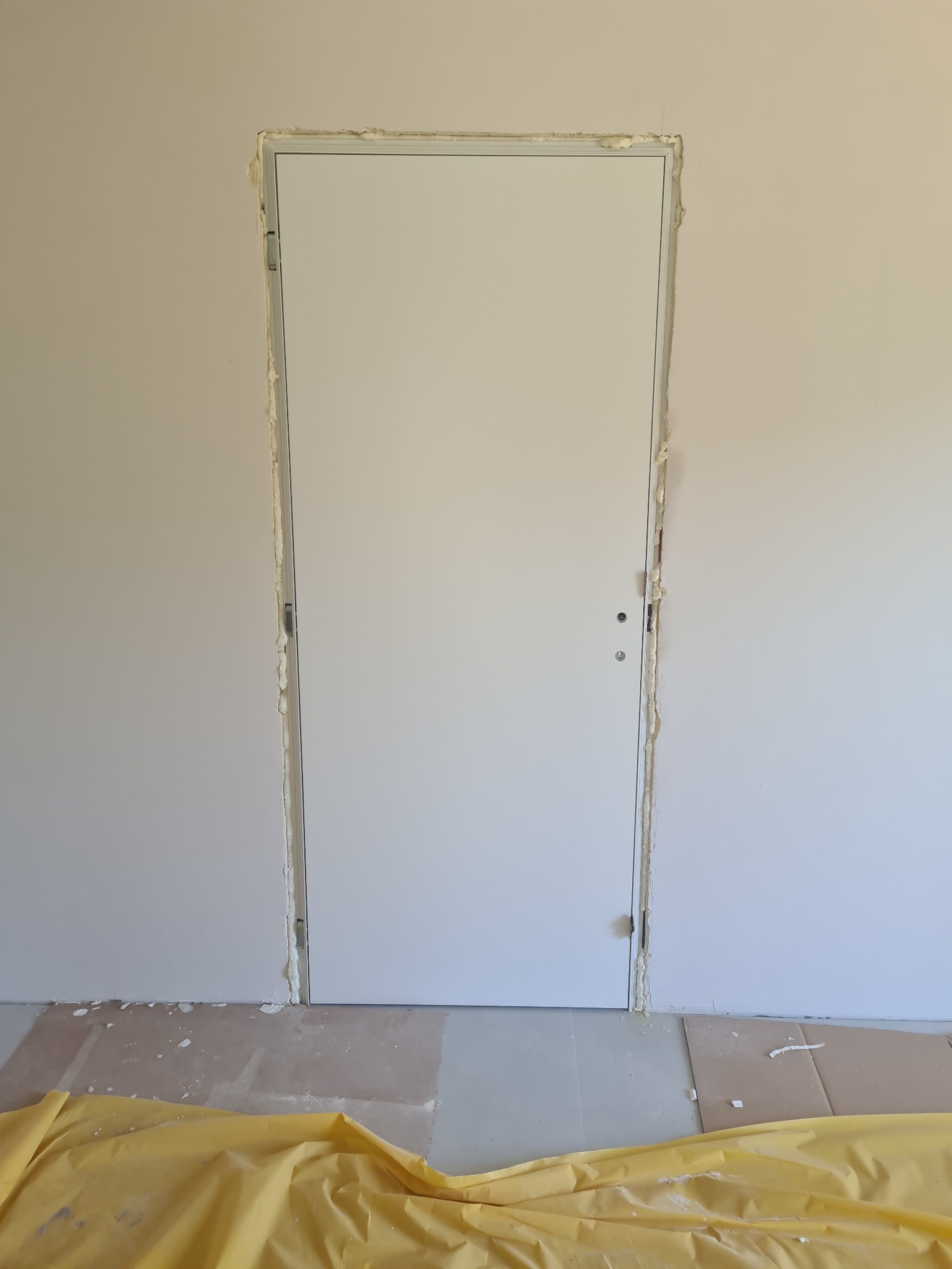 Białe drzwi wewnętrzne w trakcie osadzania w ościeżnicy, widoczna piana montażowa i zabezpieczenie podłogi folią. Ujęcie z bliska, skupione na detalu montażu.