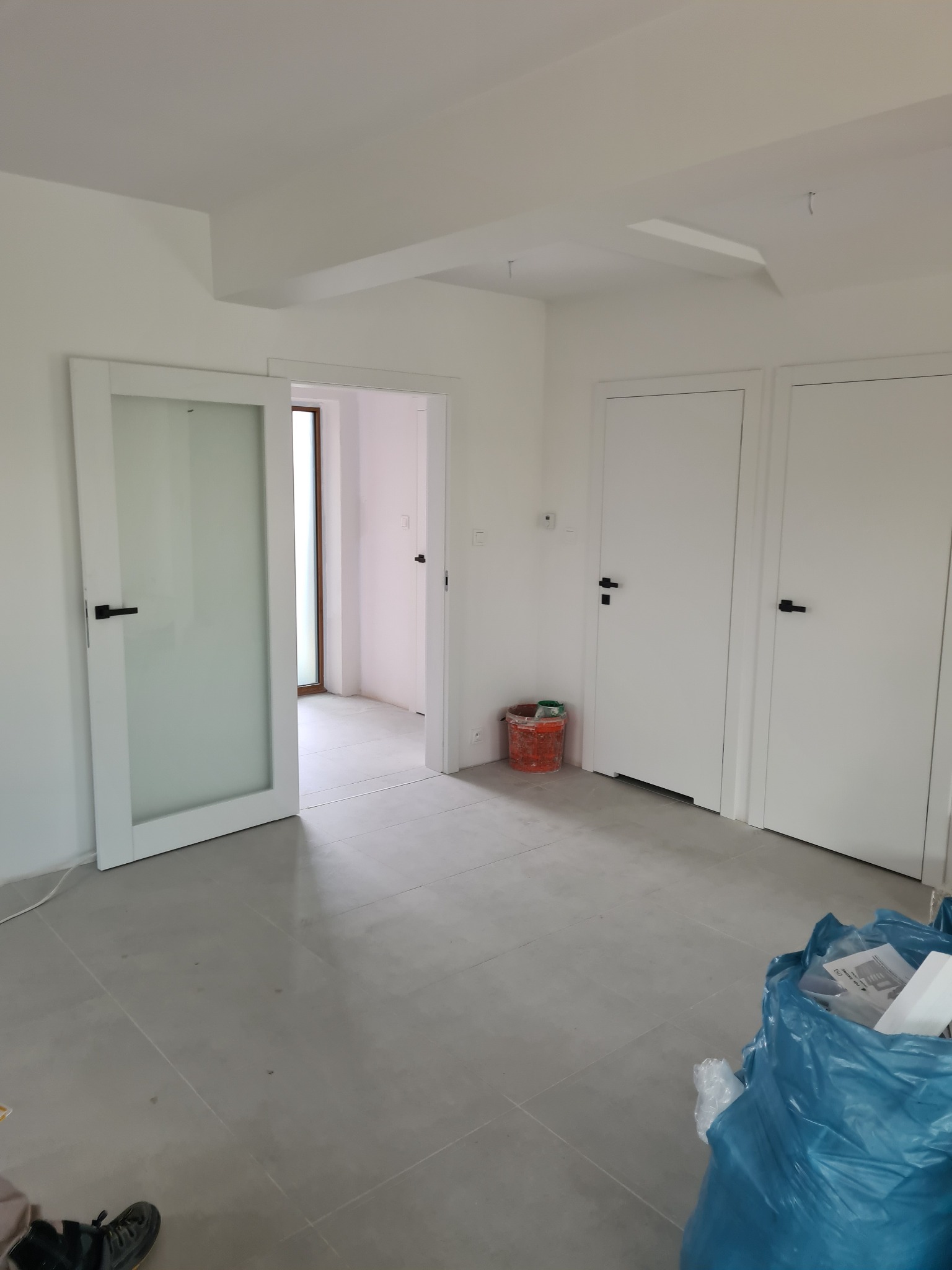 Jasne wnętrze po remoncie z trzema białymi drzwiami z czarnymi klamkami. Jedne drzwi otwarte, widoczny fragment okna w głębi. Na podłodze szare płytki.