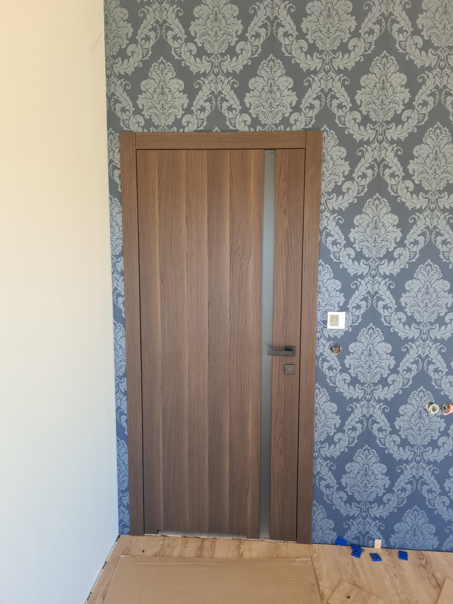 Zamontowane, nowoczesne drzwi wewnętrzne z przeszkleniem, na tle ściany z dekoracyjną tapetą w odcieniach szarości i bieli. Widoczne elementy wykończeniowe podłogi.