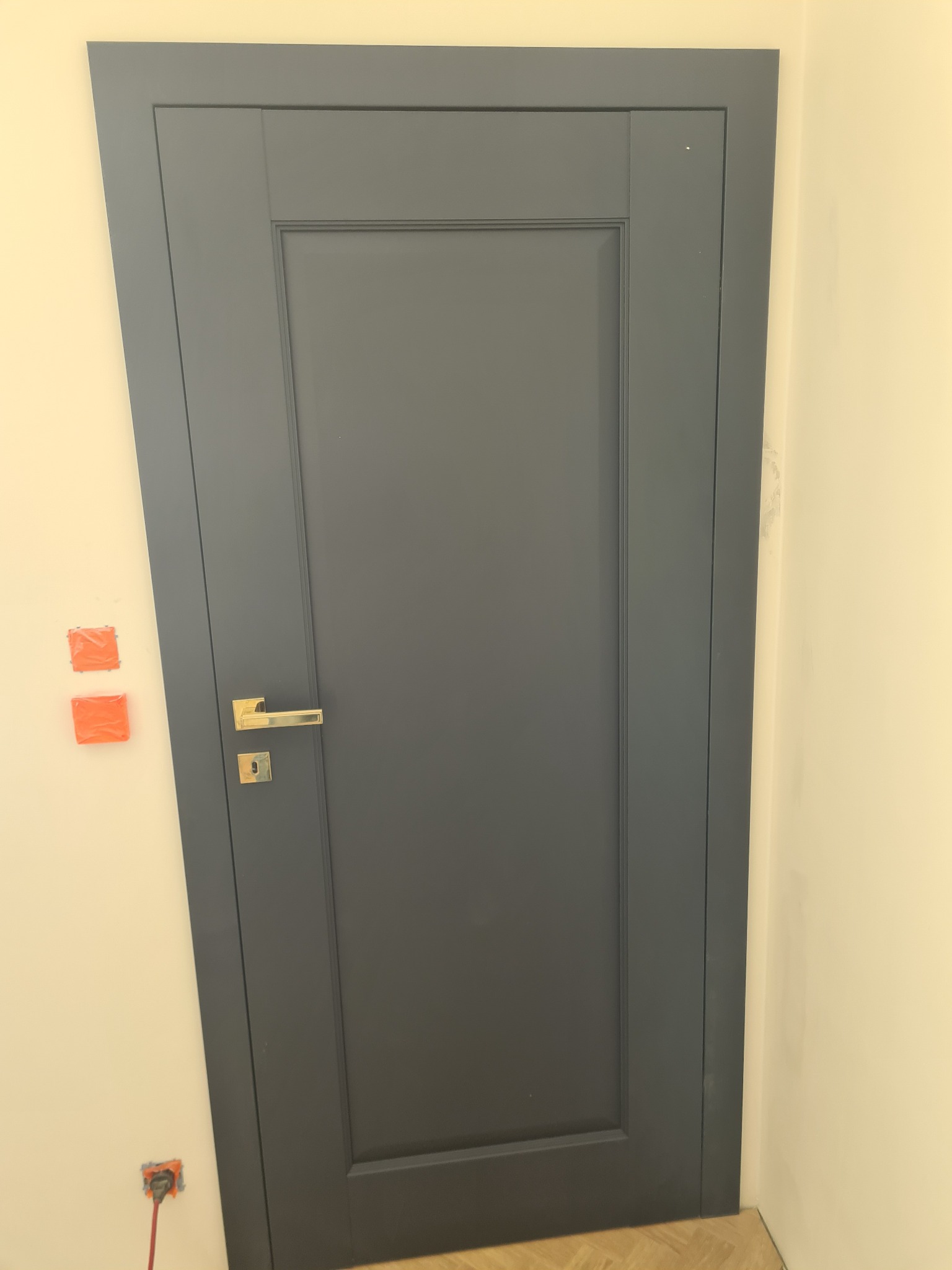 Zamknięte, grafitowe drzwi wewnętrzne z złotą klamką, widoczne pomarańczowe zabezpieczenia gniazdek elektrycznych na ścianie. Minimalistyczny design.
