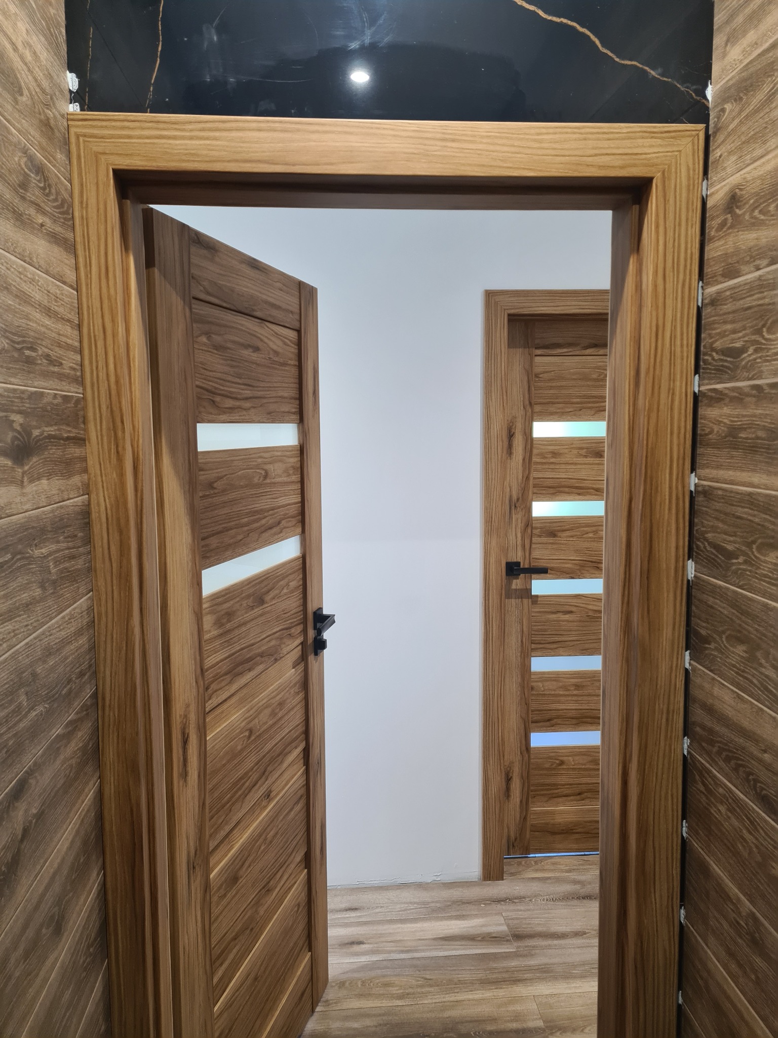 Dwa drewniane skrzydła drzwiowe z przeszkleniami, jedno otwarte, drugie zamknięte. Widoczne ościeżnice i fragment ściany z drewnopodobnymi panelami. Nowoczesny design.