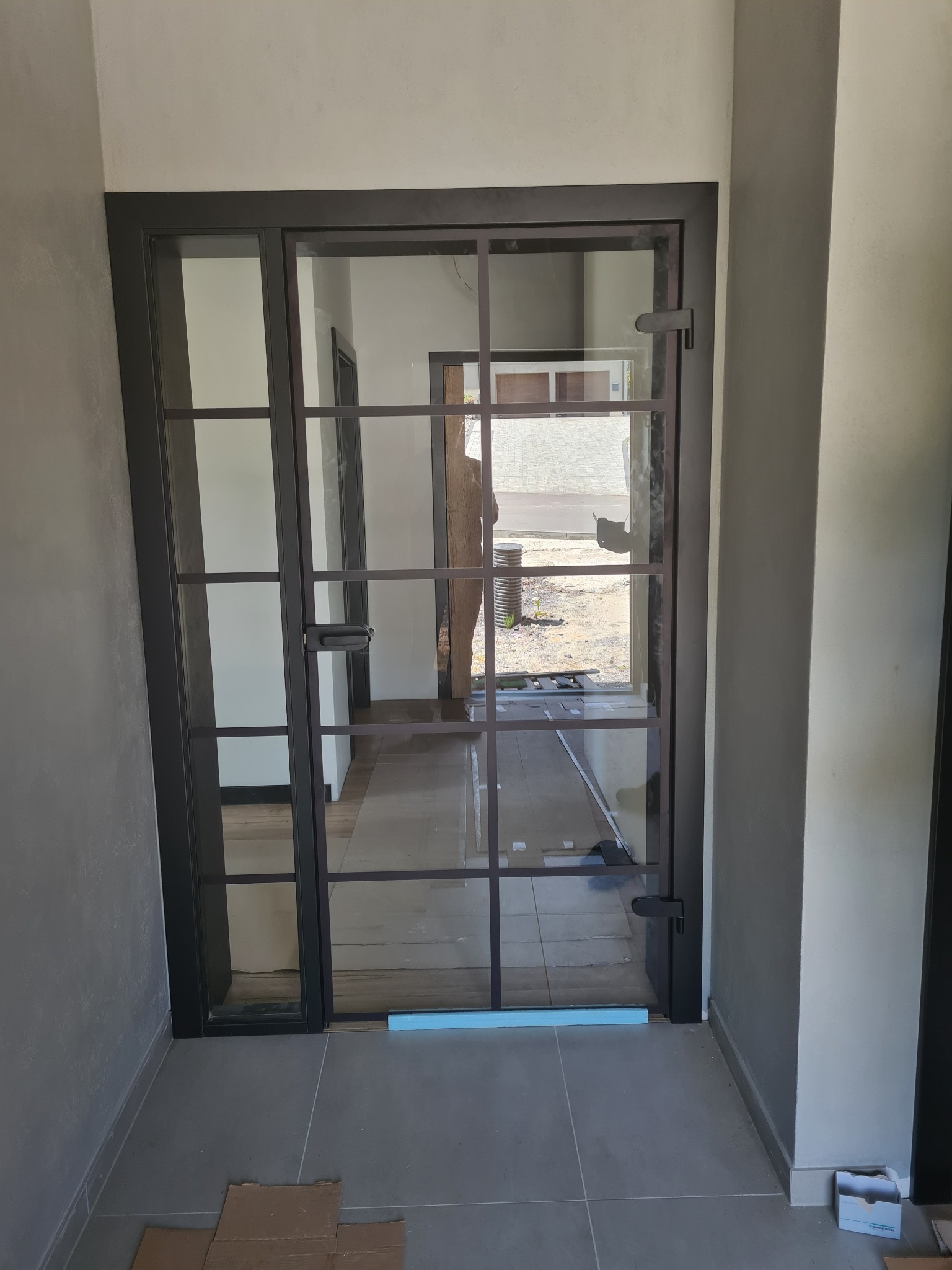 Nowoczesne, przeszklone drzwi wewnętrzne z czarną ramą i podziałami, widoczne z bliska. W tle widoczne kolejne pomieszczenia i fragment ulicy.