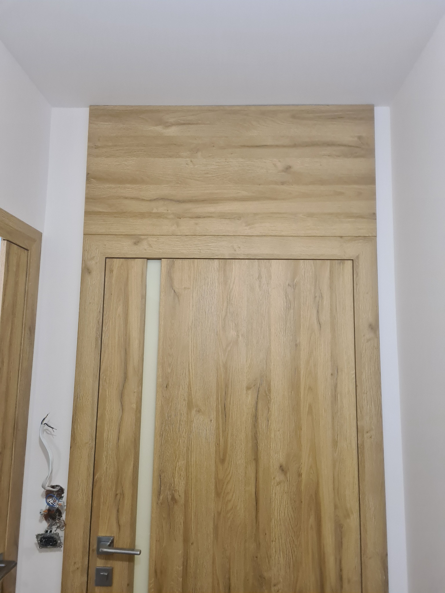 Zamknięte, drewniane drzwi wewnętrzne z mleczną szybą, dopasowane do obudowy nadproża. Po lewej widoczna instalacja elektryczna przed wykończeniem.
