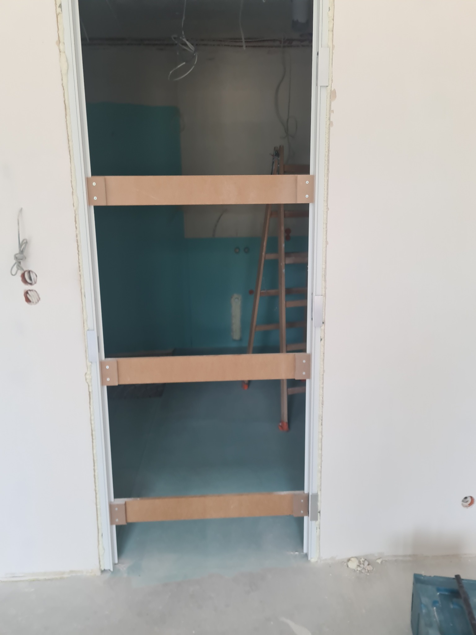 Ościeżnica drzwiowa zabezpieczona drewnianymi rozpórkami podczas montażu. W tle widoczna drabina i fragment ściany w kolorze morskim. Surowy stan wykończenia.