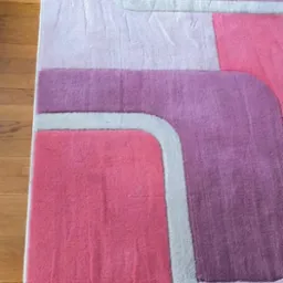 Fragment puszystego dywanu z geometrycznym wzorem w odcieniach różu, fioletu i bieli, leżącego na drewnianej podłodze