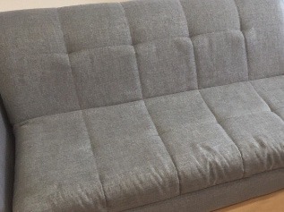 Szara, tekstylna kanapa z wyraźnymi, kwadratowymi przeszyciami, widoczna z bliska, z lekkimi śladami użytkowania.