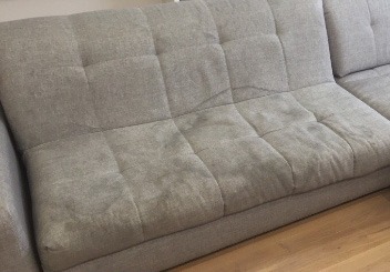 Szara, modułowa kanapa z widocznymi, ciemniejszymi plamami na oparciu i siedzisku, sugerującymi zabrudzenia przed czyszczeniem.